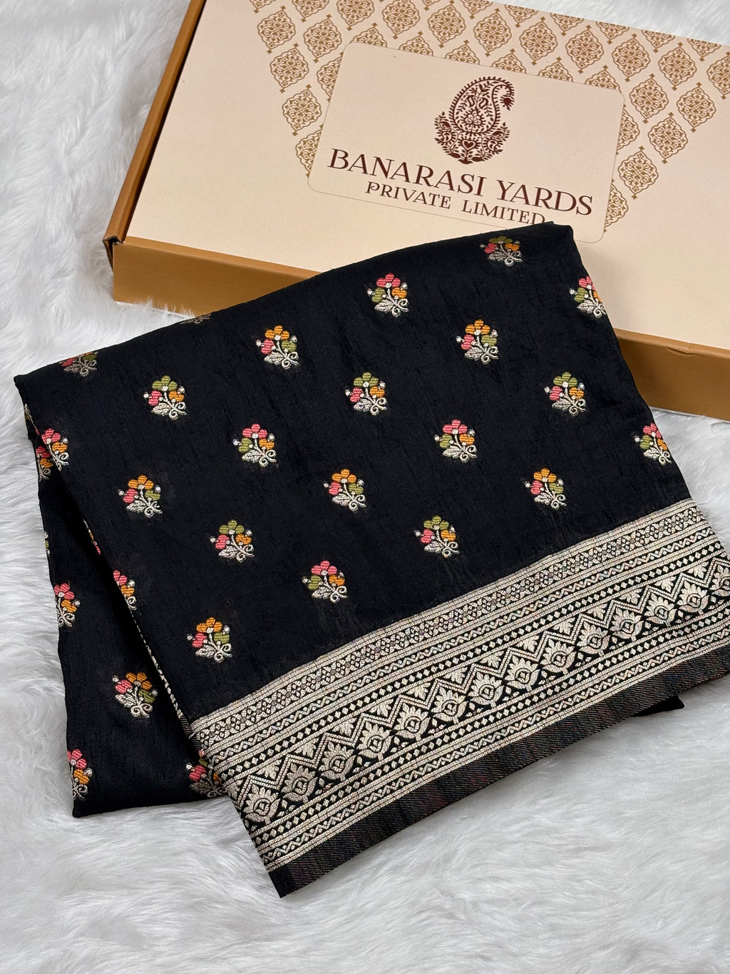 A folded black Banarasi saree with small multicolor floral motifs and a wide beige-and-black patterned border, displayed on a white faux-fur surface beside a beige Banarasi Yards box.