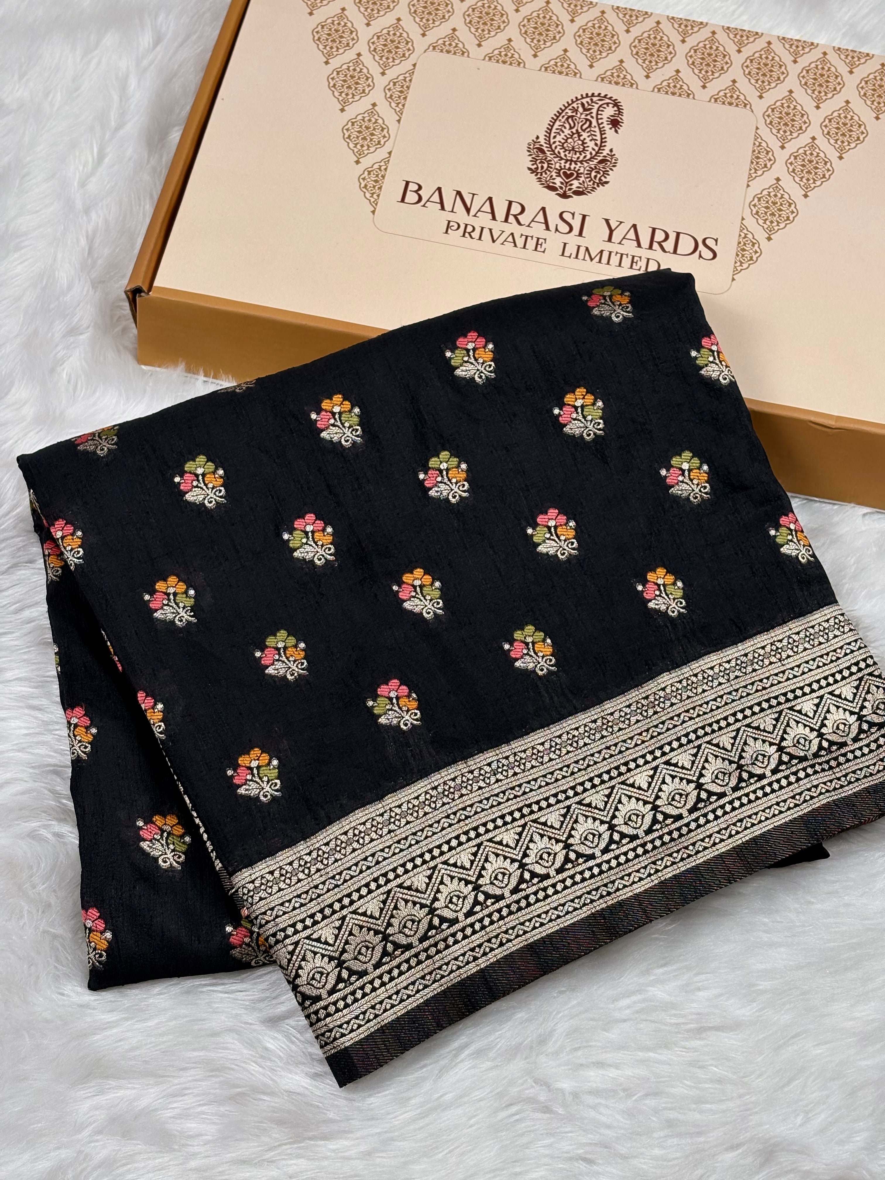 A folded black Banarasi saree with small multicolor floral motifs and a wide beige-and-black patterned border, displayed on a white faux-fur surface beside a beige Banarasi Yards box.