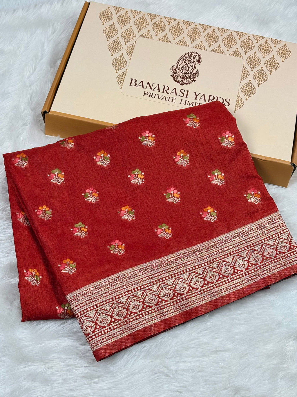 A folded red Banarasi Yards silk saree with small floral motifs and a decorative border, displayed next to its branded packaging box on a white faux fur surface.