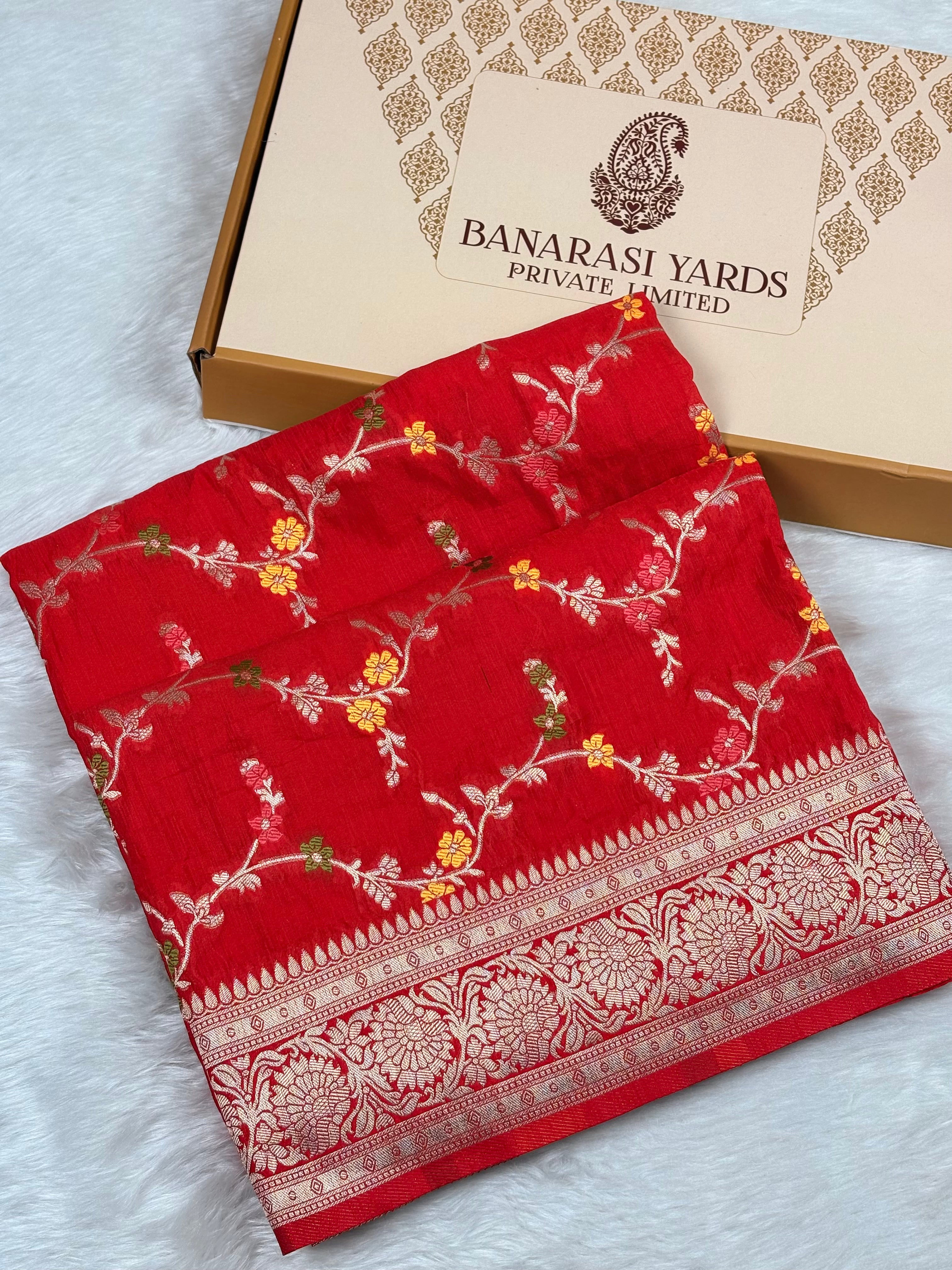 A folded red Banarasi saree with floral weaving and a decorative border, placed in front of a Banarasi Yards Private Limited box on a white textured surface.