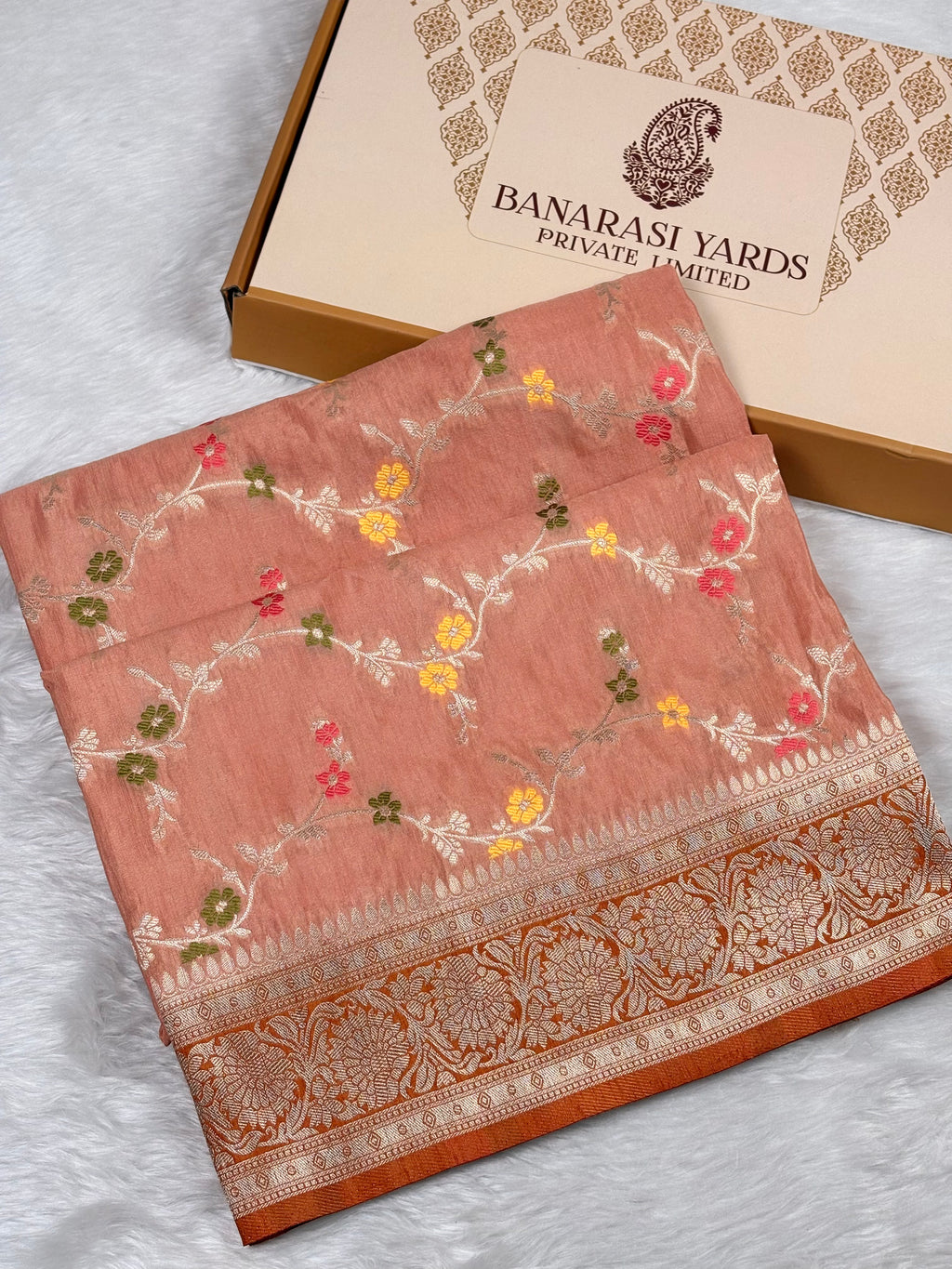 A folded peach-pink Banarasi saree with floral weaving and a decorative border, displayed next to a Banarasi Yards box on a white textured surface.