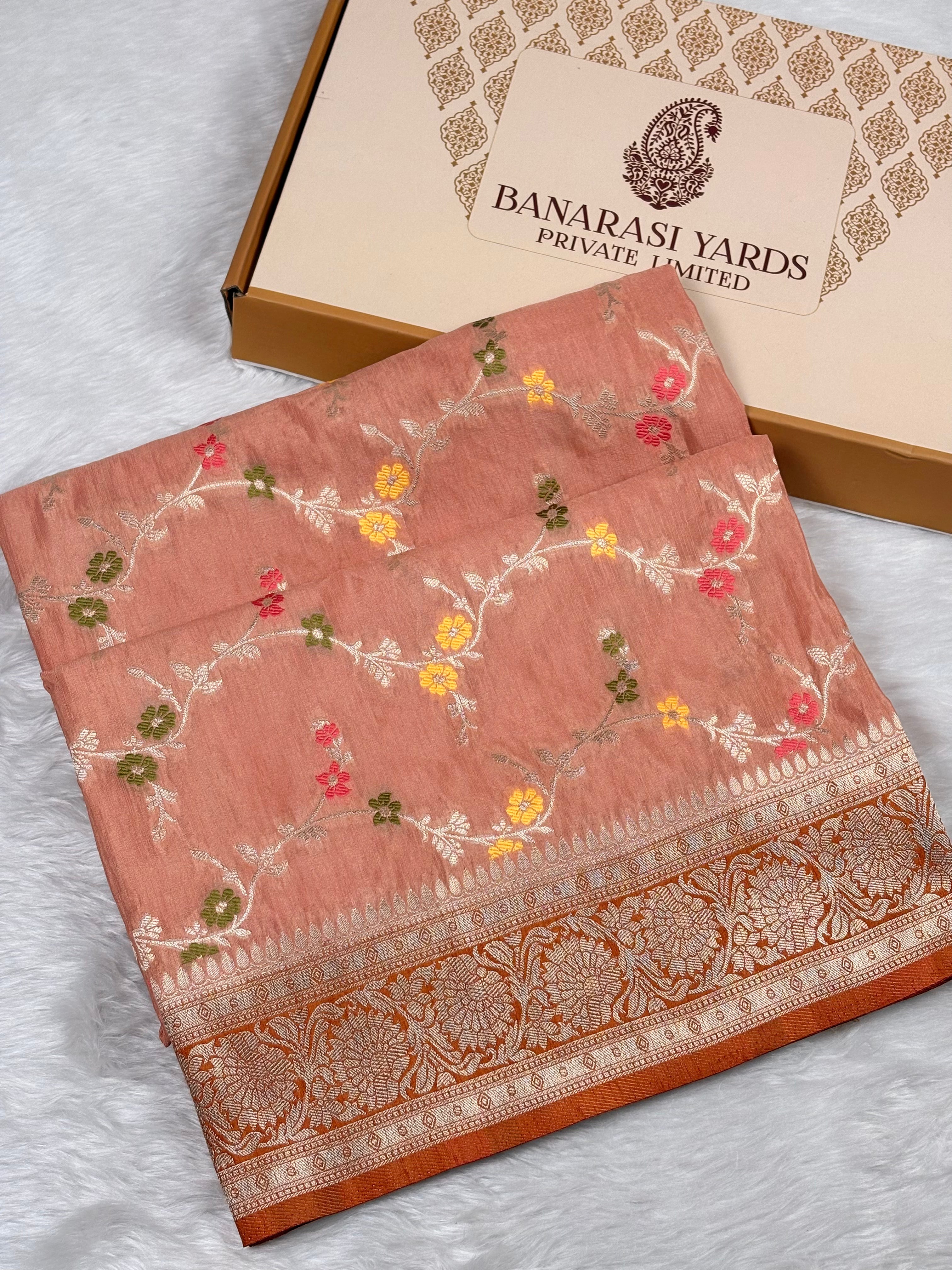 A folded peach-pink Banarasi saree with floral weaving and a decorative border, displayed next to a Banarasi Yards box on a white textured surface.
