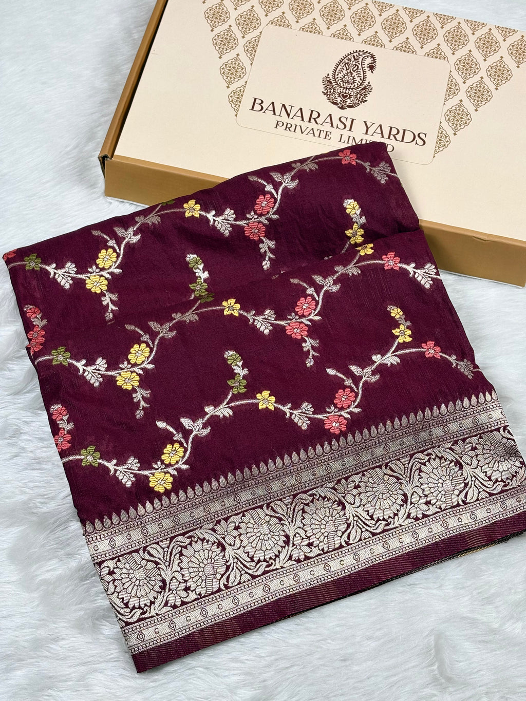 A folded deep maroon saree with intricate floral weaving and a wide metallic border, displayed next to its packaging box labeled 'BANARASI YARDS PRIVATE LIMITED' on a white faux fur surface.