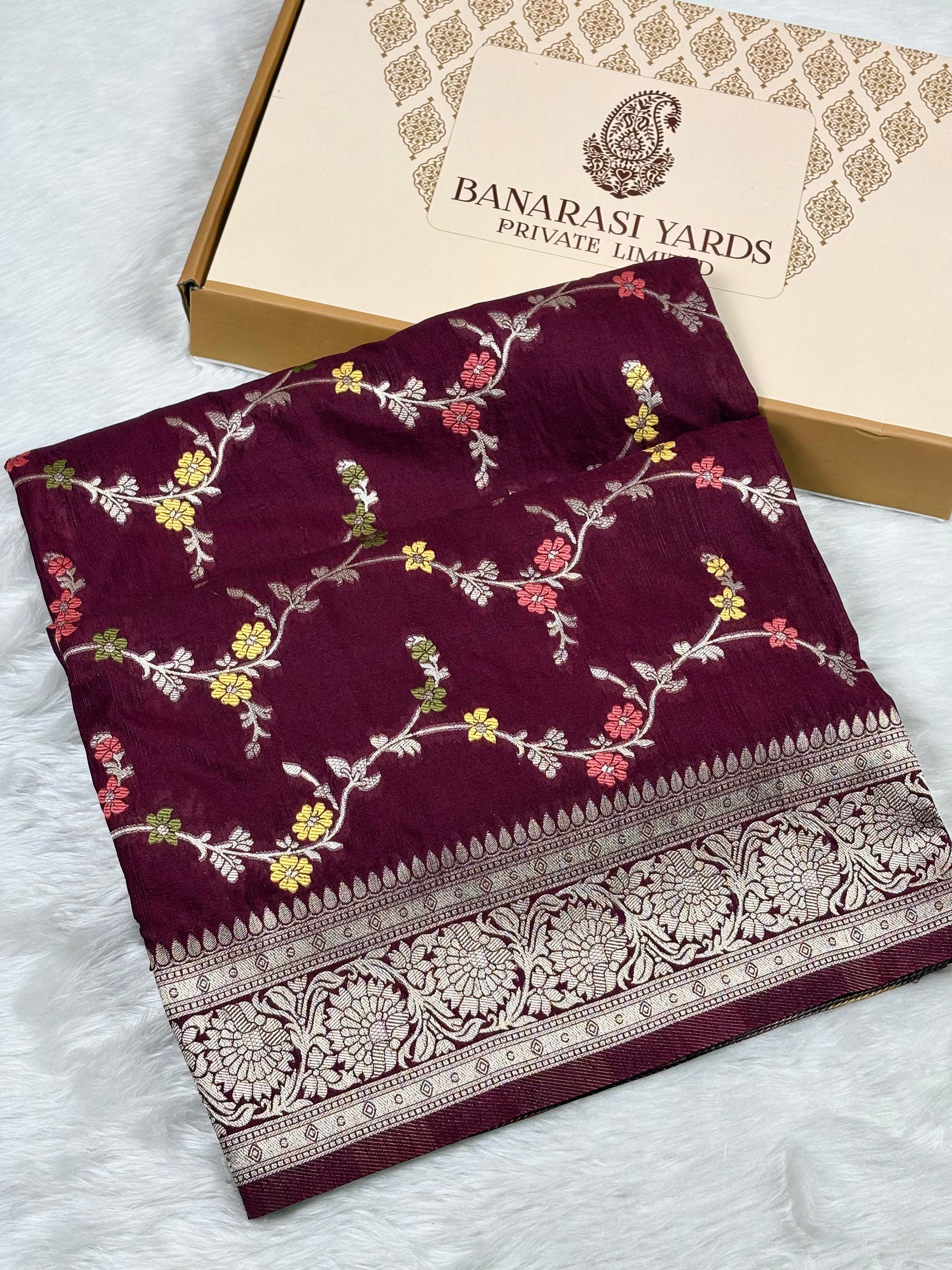 A folded deep maroon saree with intricate floral weaving and a wide metallic border, displayed next to its packaging box labeled 'BANARASI YARDS PRIVATE LIMITED' on a white faux fur surface.