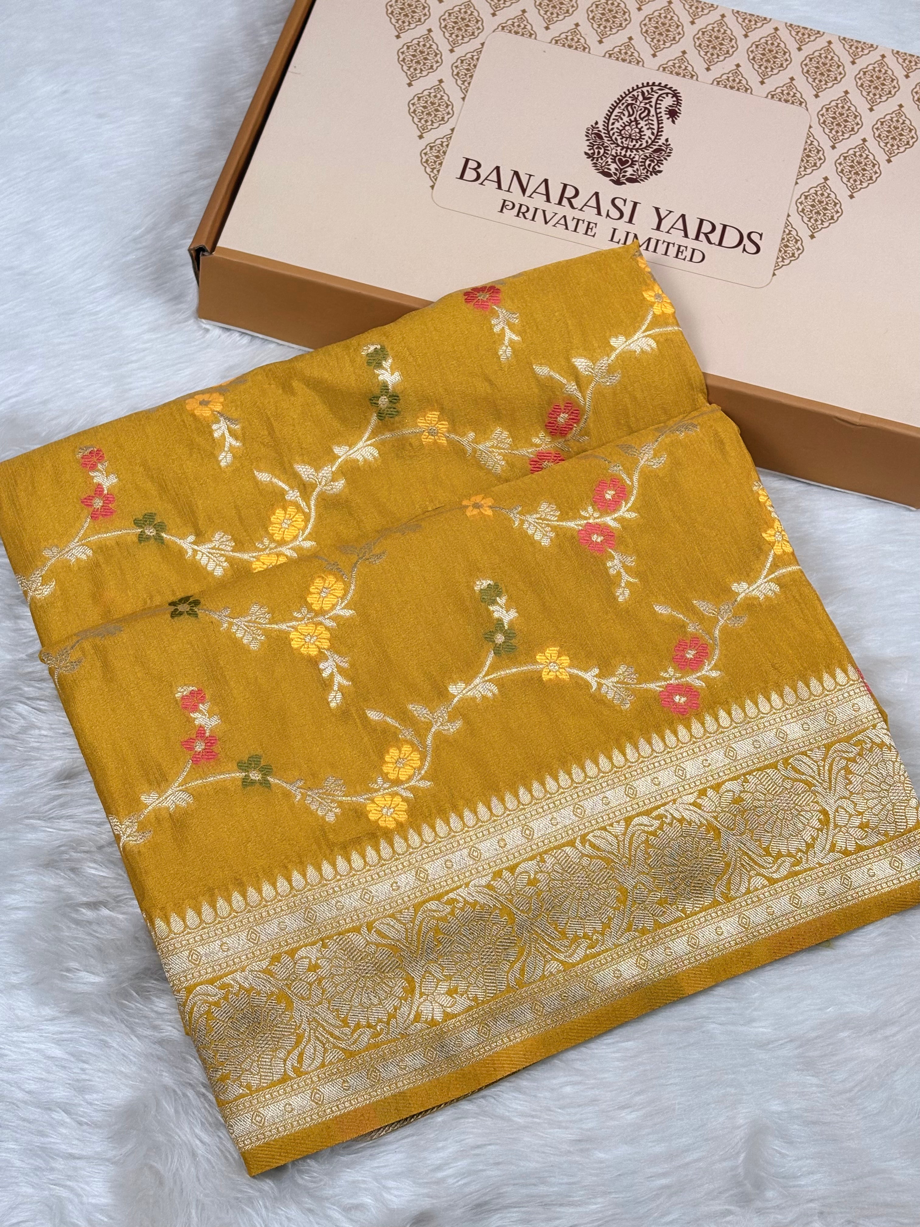 A folded mustard yellow Banarasi saree with intricate gold zari weaving and floral motifs, displayed next to its branded packaging box on a white faux fur surface.