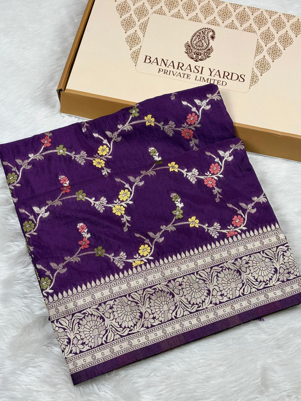 A folded purple Banarasi saree with intricate gold floral weaving and a wide gold border, displayed next to its beige 'Banarasi Yards Private Limited' box on a white faux fur surface.