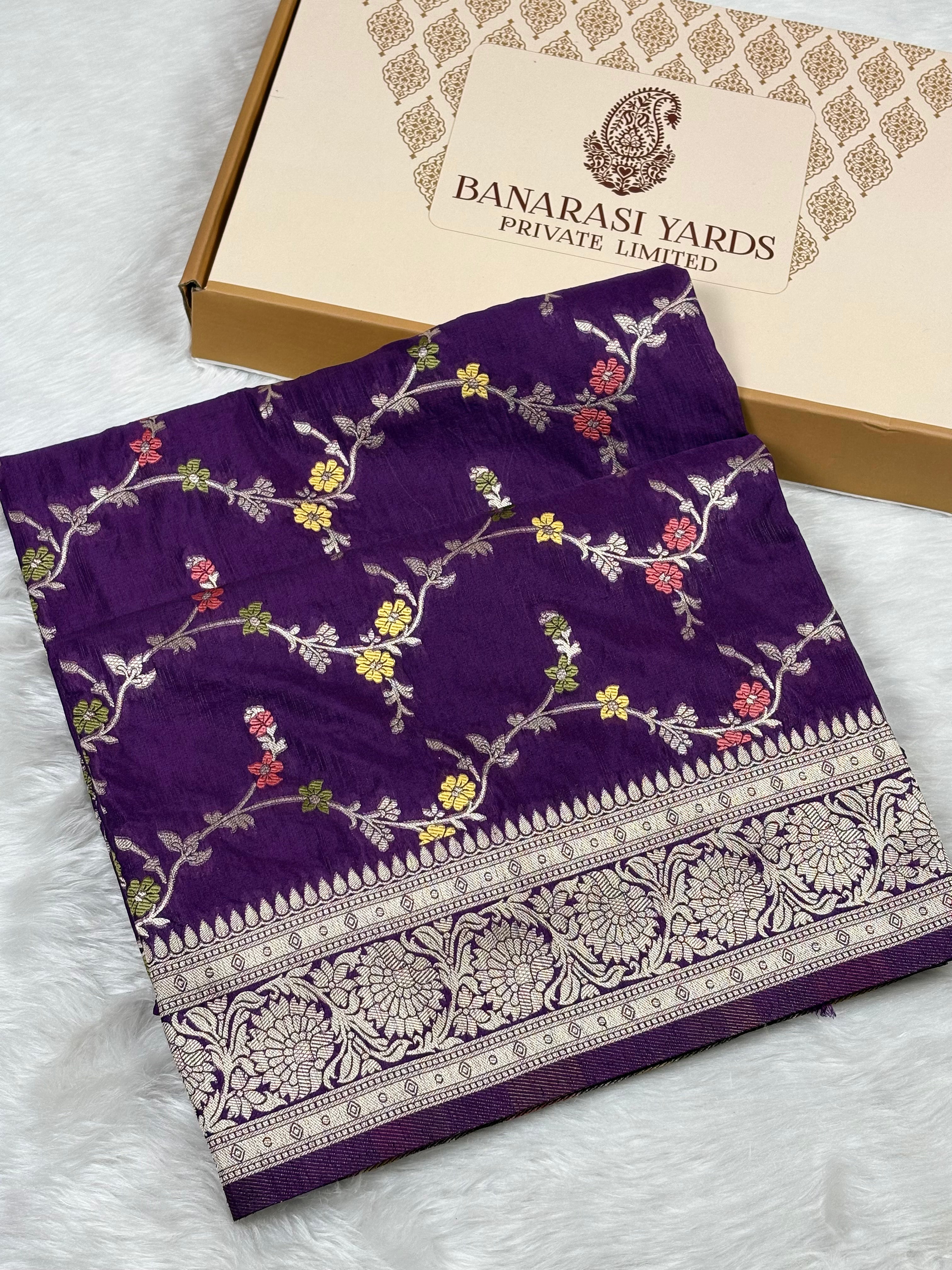 A folded purple Banarasi saree with intricate gold floral weaving and a wide gold border, displayed next to its beige 'Banarasi Yards Private Limited' box on a white faux fur surface.