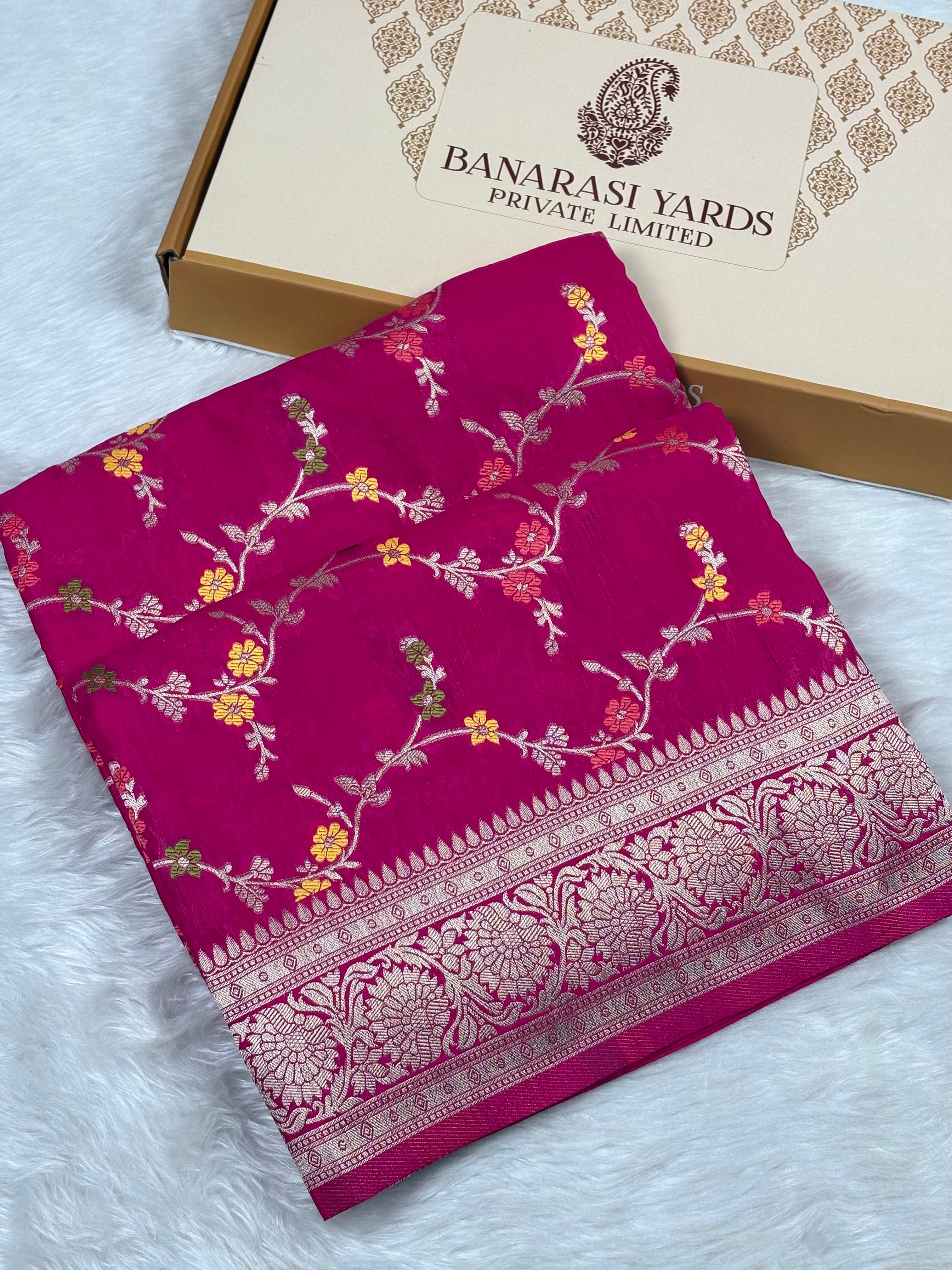 A folded magenta pink Banarasi silk saree with intricate gold floral weaving and a decorative border, displayed next to its 'Banarasi Yards Private Limited' box on a white faux fur surface.
