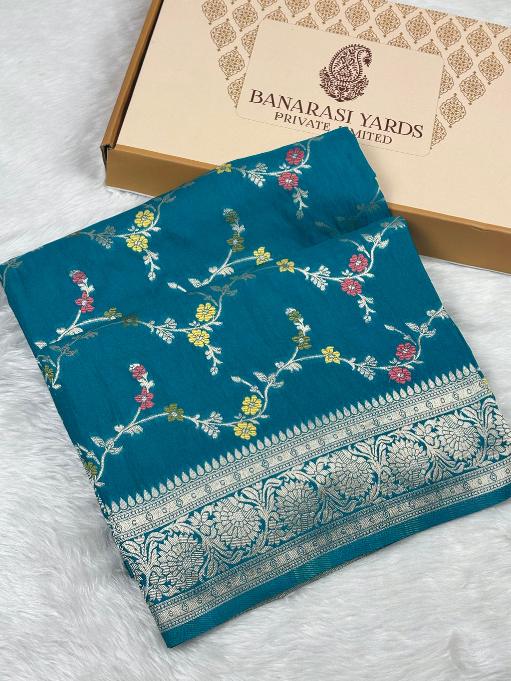A folded teal blue Banarasi saree with intricate floral weaving and a detailed zari border, displayed next to its cardboard packaging box on a white faux fur surface.