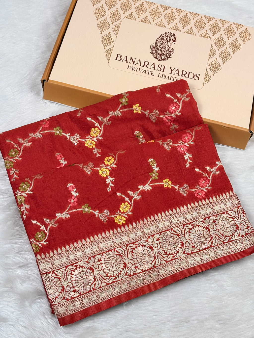 A folded red Banarasi silk saree with intricate gold, pink, and green floral mina and a wide gold zari border, displayed next to its beige Banarasi Yards Private Limited box on a white faux fur surface.
