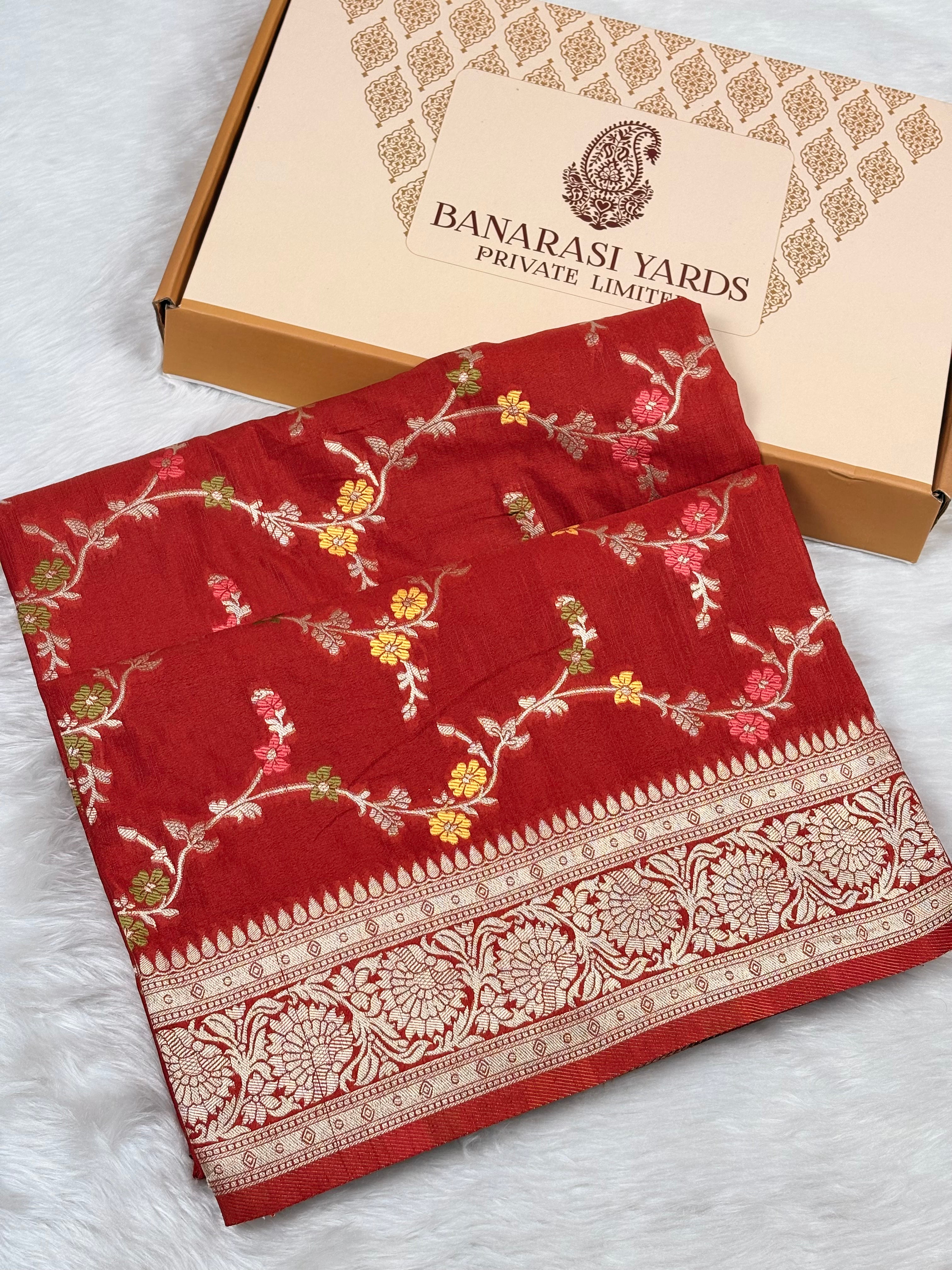 A folded red Banarasi silk saree with intricate gold, pink, and green floral mina and a wide gold zari border, displayed next to its beige Banarasi Yards Private Limited box on a white faux fur surface.
