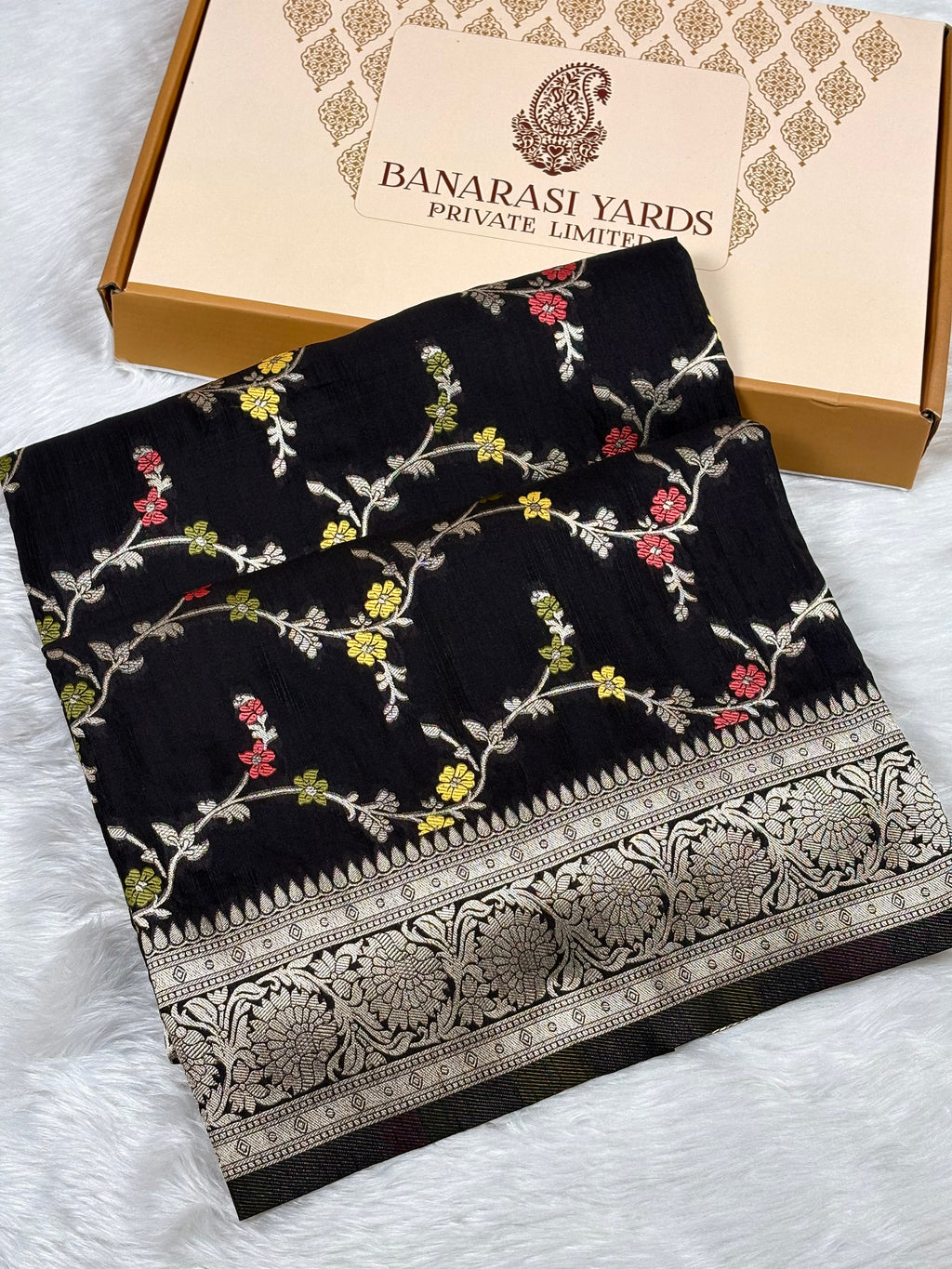 Black Banarasi silk saree with colorful floral mina and a gold zari border, displayed next to its beige Banarasi Yards Private Limited box on a white faux fur surface.