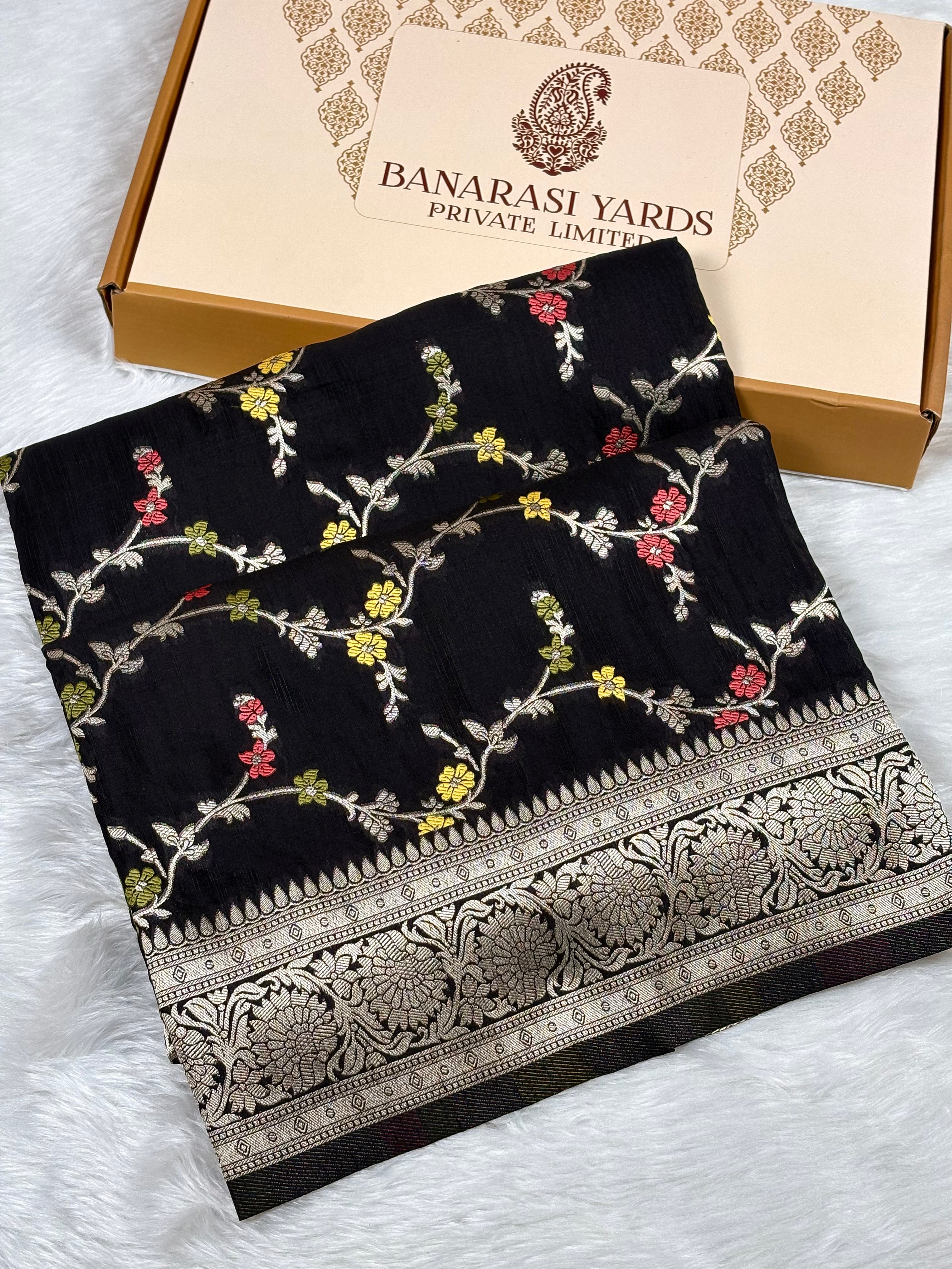 Black Banarasi silk saree with colorful floral mina and a gold zari border, displayed next to its beige Banarasi Yards Private Limited box on a white faux fur surface.