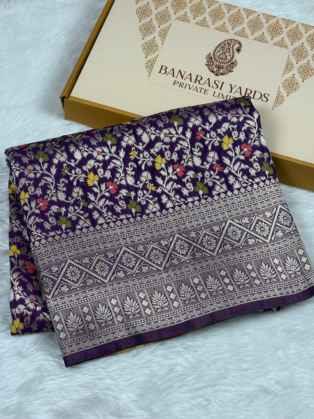 A folded purple Banarasi silk saree with intricate floral and geometric patterns, displayed next to its branded packaging box on a white faux fur surface.