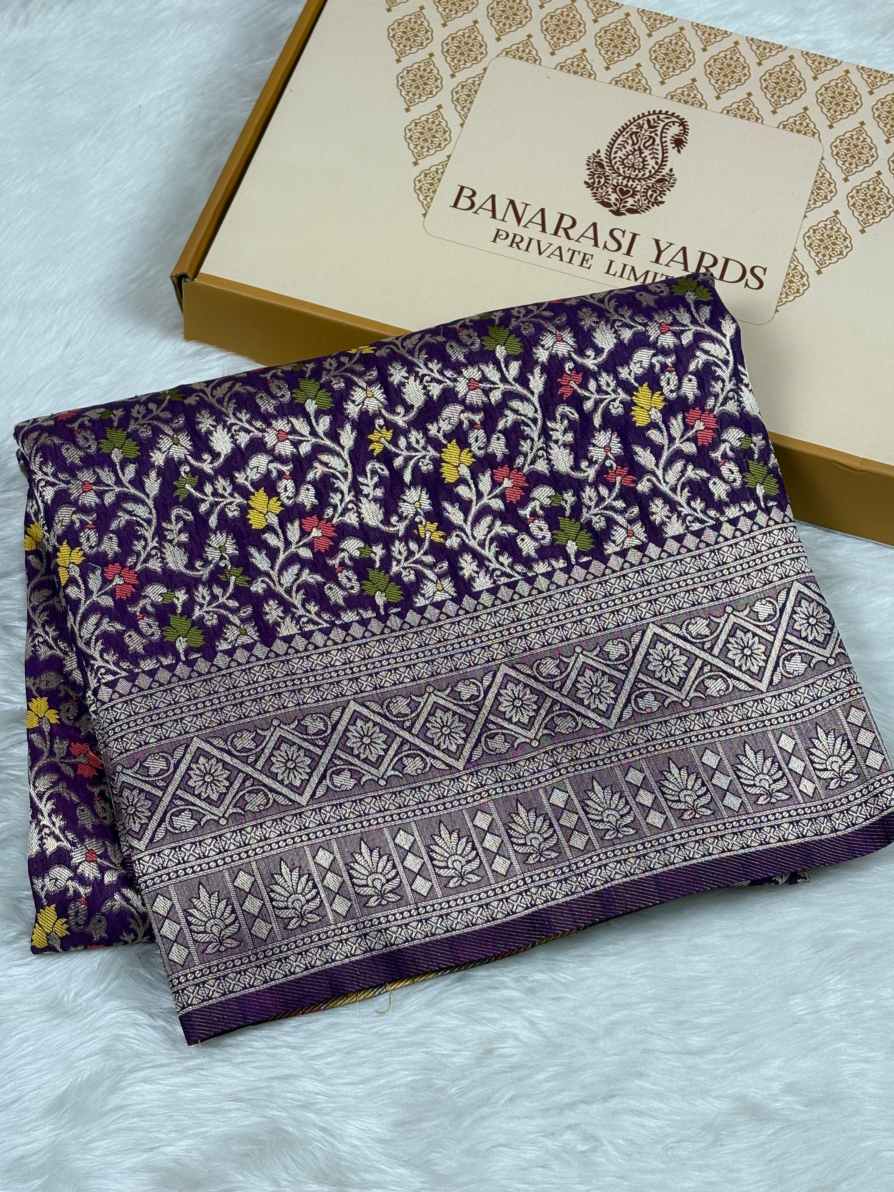 A folded purple Banarasi silk saree with intricate floral and geometric patterns, displayed next to its branded packaging box on a white faux fur surface.