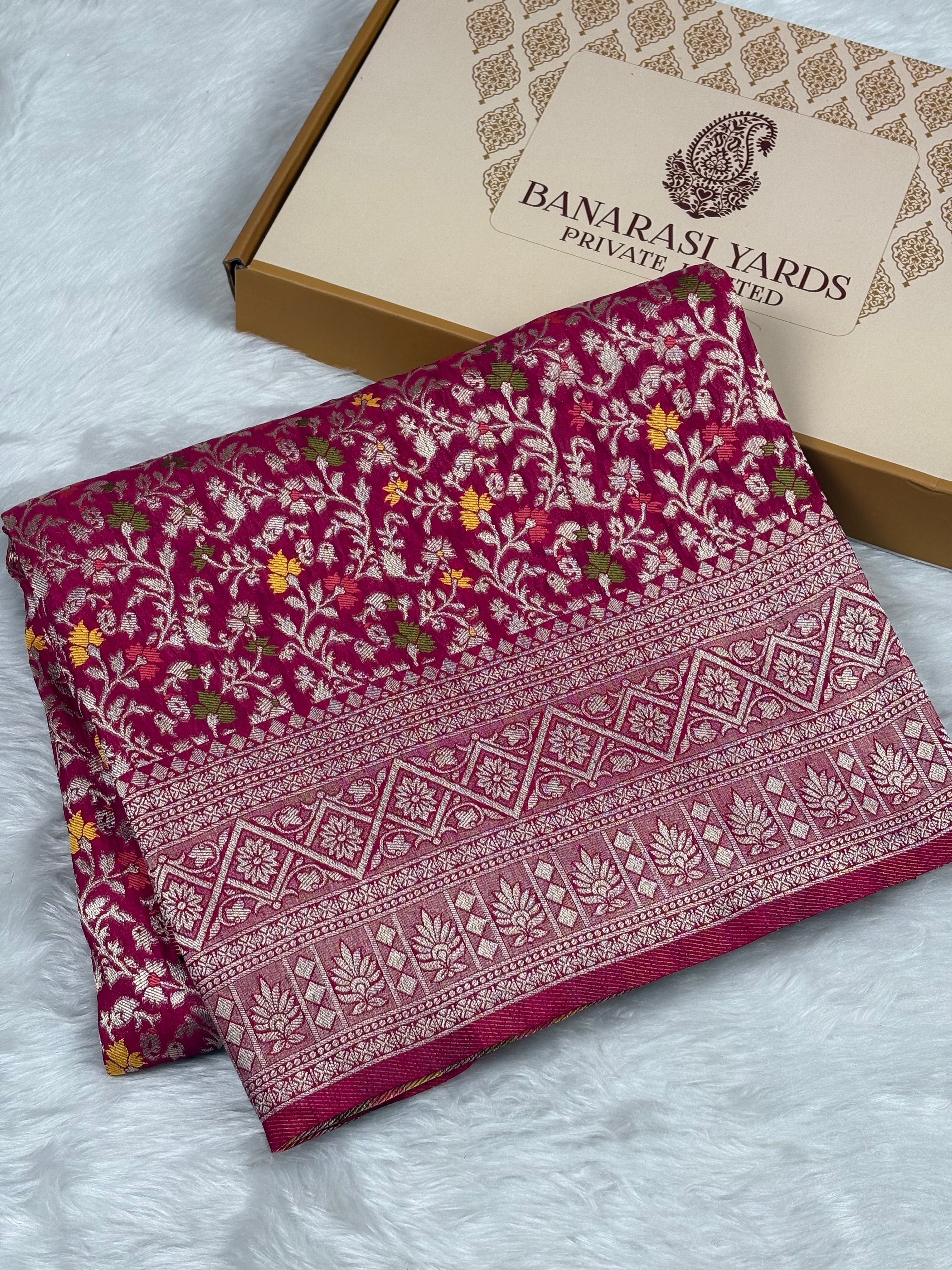 A folded deep magenta Banarasi silk saree with intricate floral zari patterns, displayed next to its packaging box on a white faux fur surface.