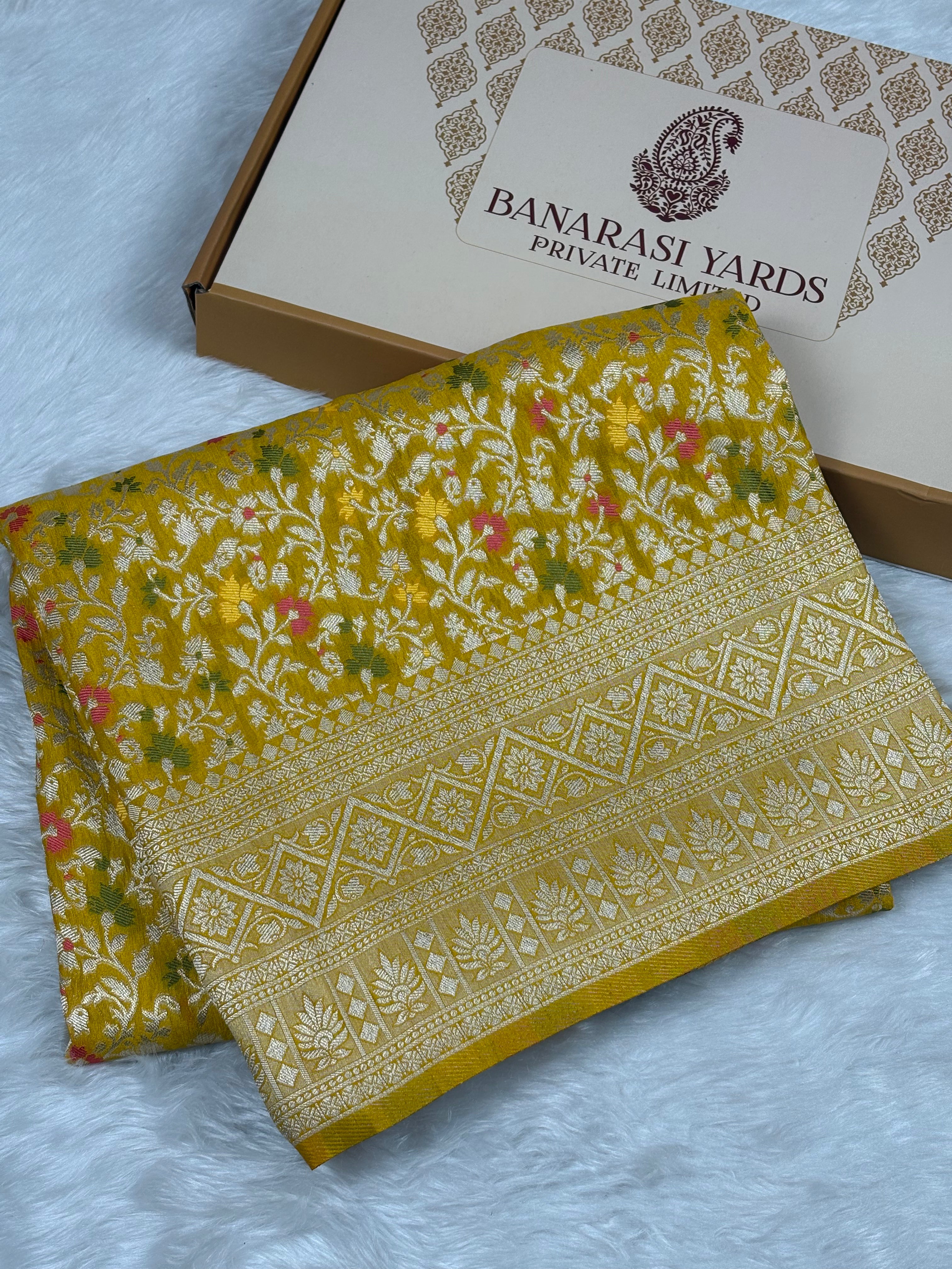 A folded mustard yellow Banarasi silk saree with intricate zari floral and geometric patterns, displayed next to its branded cardboard box on a white faux fur surface.