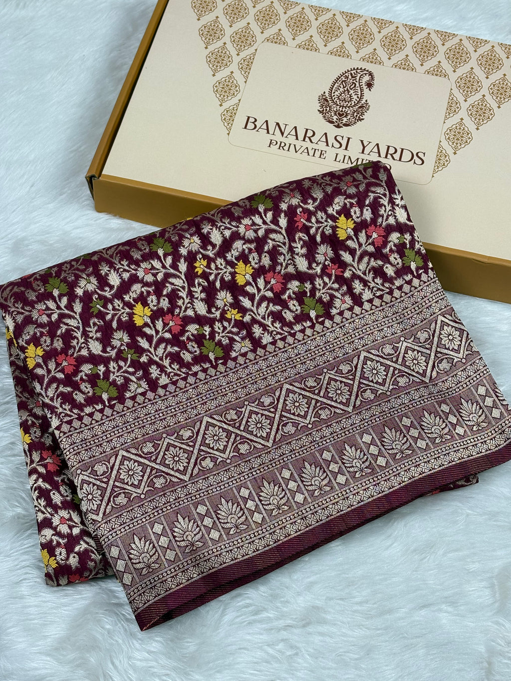 A folded dark purple Banarasi silk saree with intricate floral patterns, displayed next to Banarasi Yards gift box on a white faux fur background.