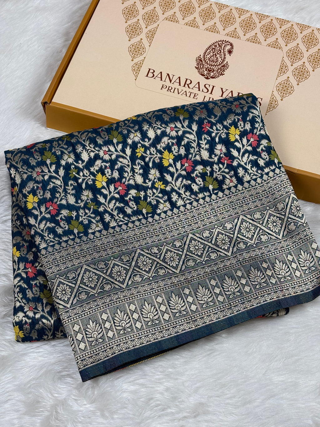 A folded navy blue Banarasi silk saree with intricate floral and geometric weaving, displayed next to its beige branded packaging box on a white faux fur surface.