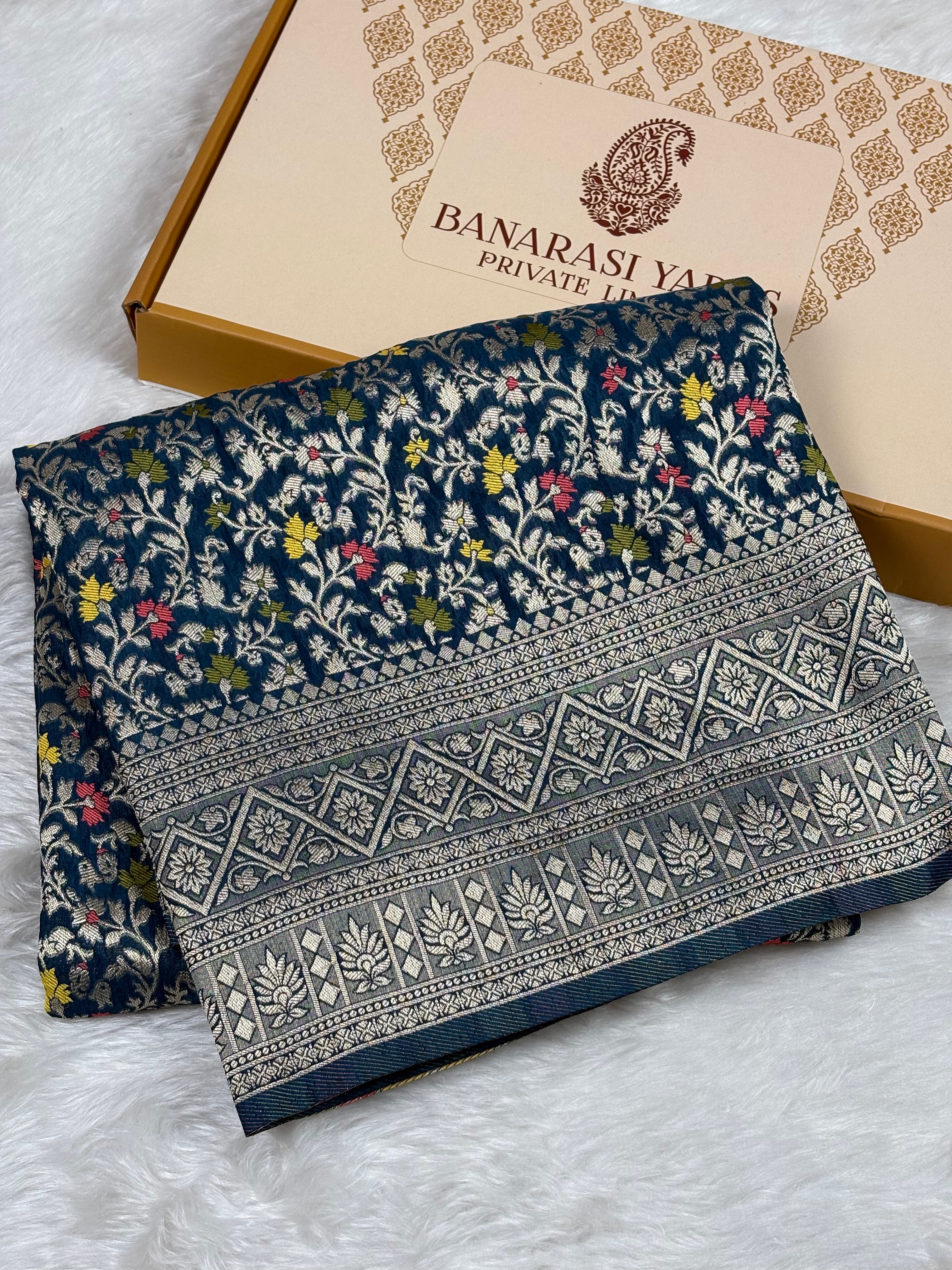 A folded navy blue Banarasi silk saree with intricate floral and geometric weaving, displayed next to its beige branded packaging box on a white faux fur surface.