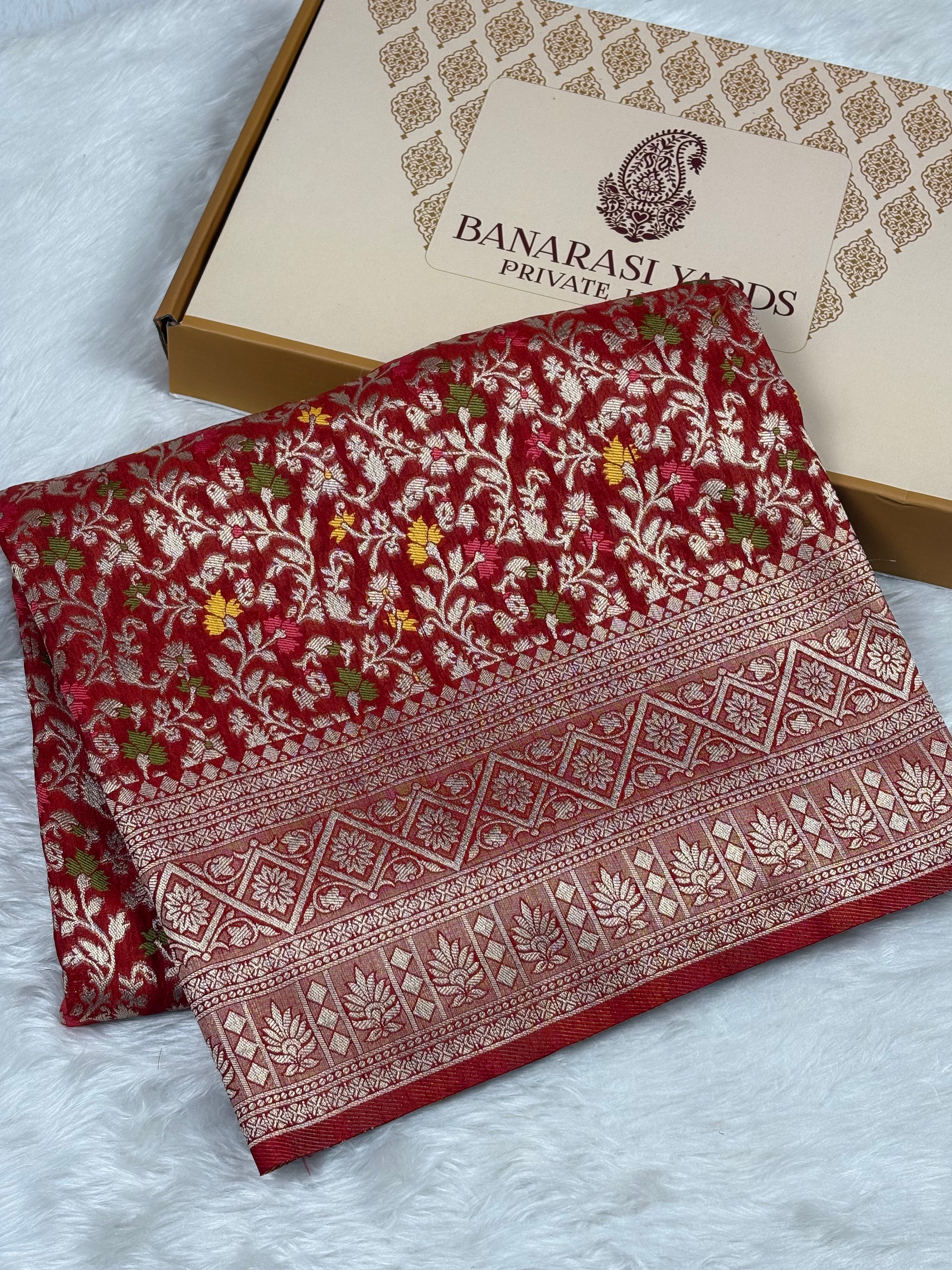 A folded red Banarasi silk saree with intricate gold floral and geometric patterns, displayed next to its beige packaging box on a white faux fur surface.