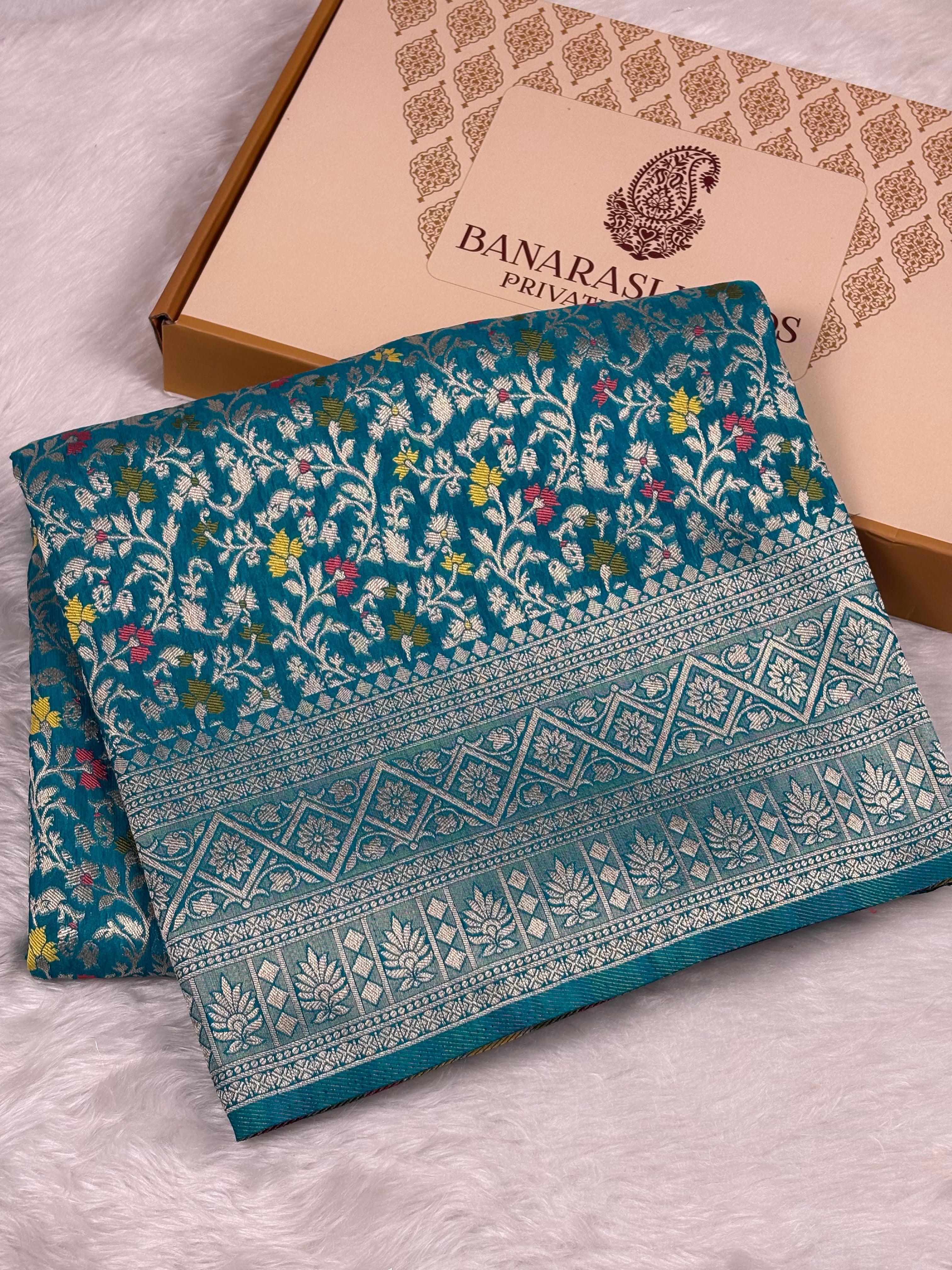 A folded teal blue Banarasi silk saree with intricate floral zari patterns and a detailed border, displayed next to its beige presentation box on a white faux fur surface.