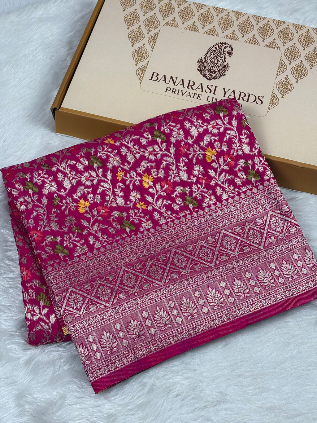 A folded magenta pink Banarasi silk saree with intricate gold and silver zari floral patterns and borders, displayed next to its beige and brown Banarasi Yards Private Limited box on a white faux fur background.