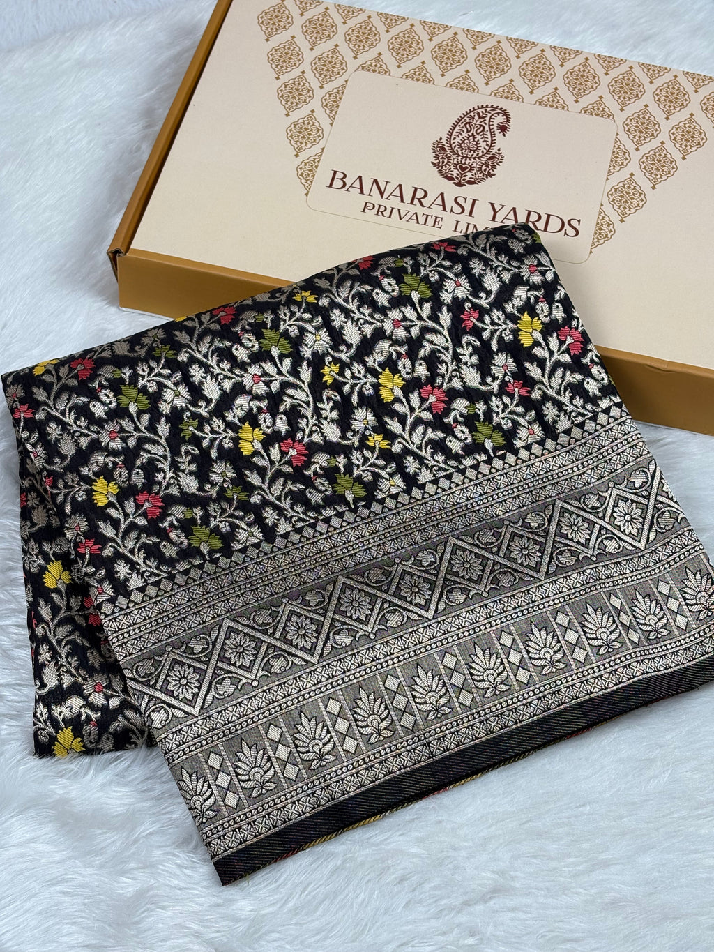 A folded black Banarasi silk saree with colorful floral mina and a detailed gold border, displayed next to its beige packaging box on a white faux fur surface. The box is branded 'BANARASI YARDS PRIVATE TRADE'.