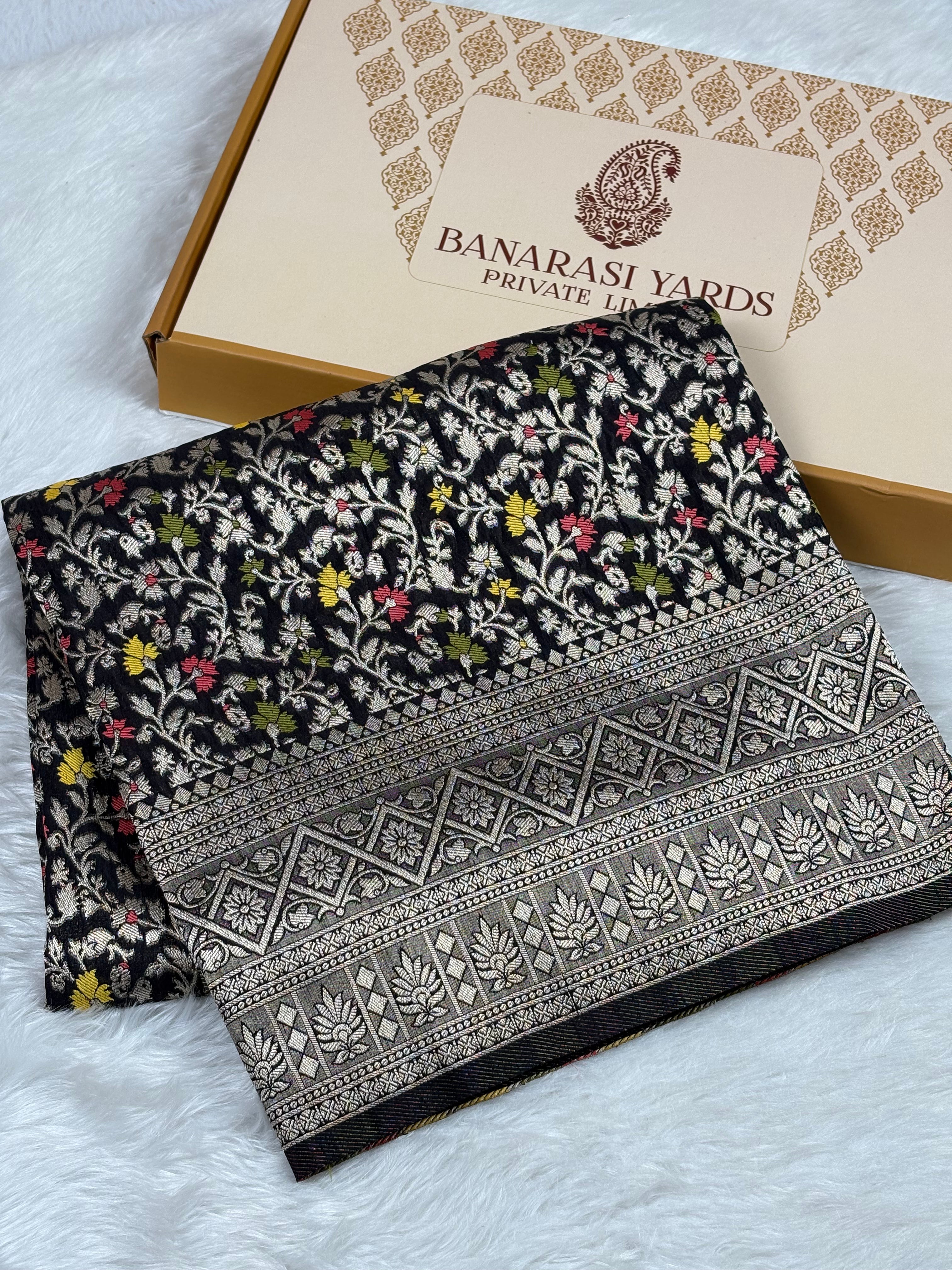 A folded black Banarasi silk saree with colorful floral mina and a detailed gold border, displayed next to its beige packaging box on a white faux fur surface. The box is branded 'BANARASI YARDS PRIVATE TRADE'.