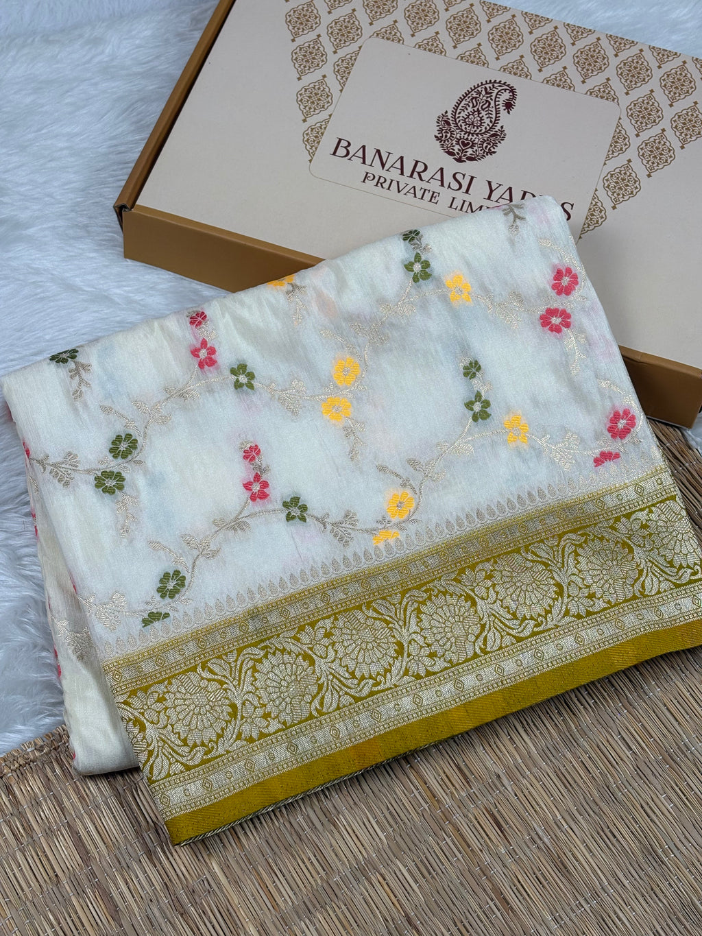 A folded white Banarasi silk saree with colorful floral mina weaving and a gold zari border, displayed next to its branded packaging box on a woven mat.