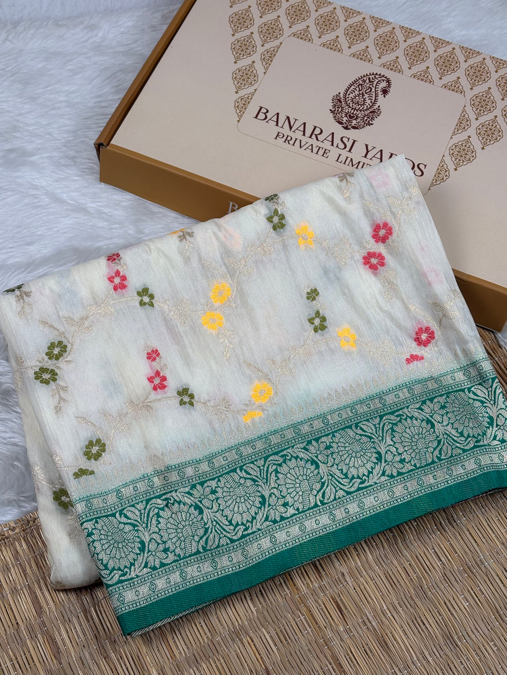 A folded white Banarasi silk saree with colorful floral mina weaving and a teal border, displayed next to its branded packaging box on a woven mat.
