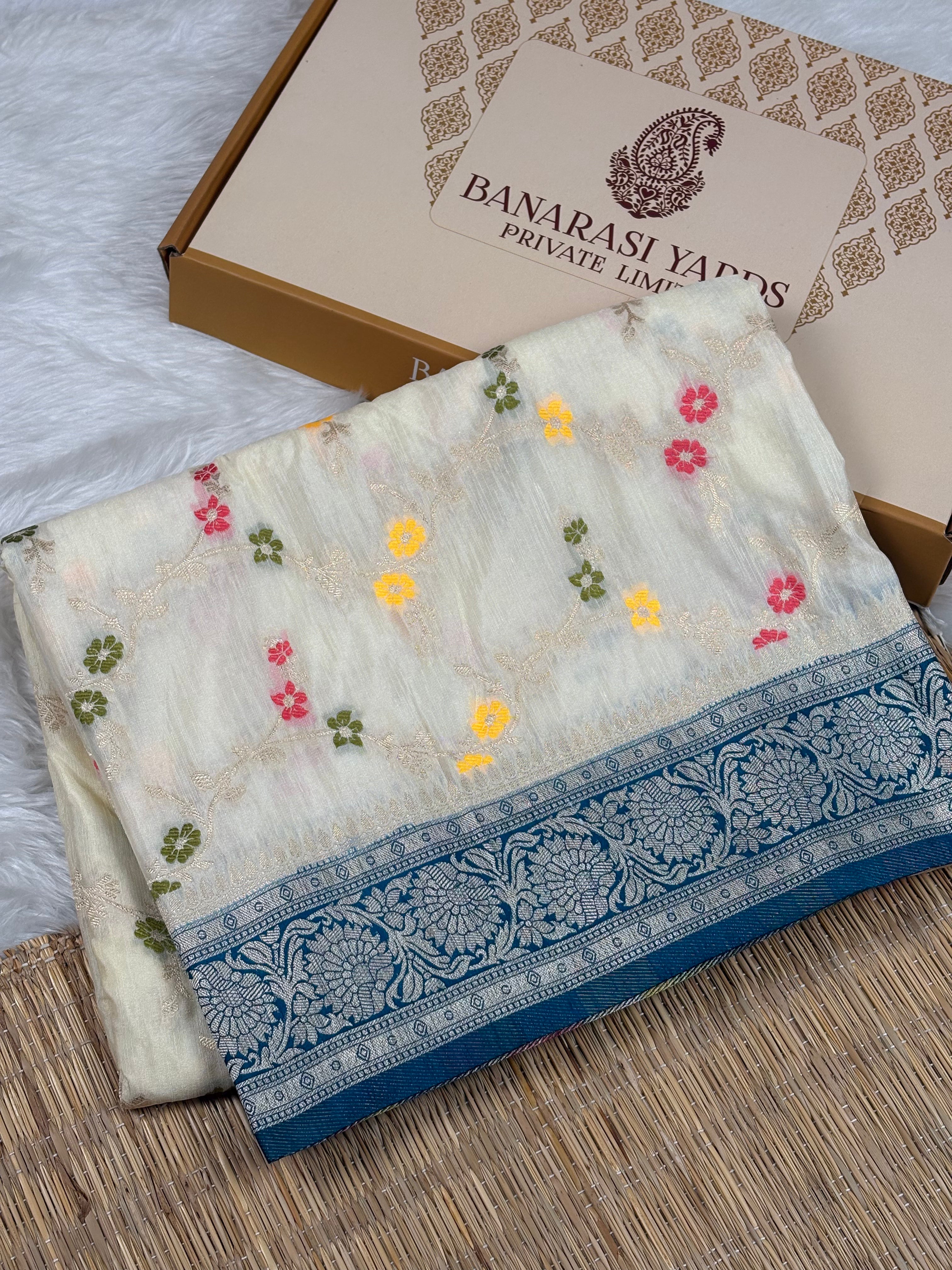 A folded cream-colored Banarasi silk saree with colorful floral mina weaving and a blue border, displayed next to its branded cardboard box on a woven mat.