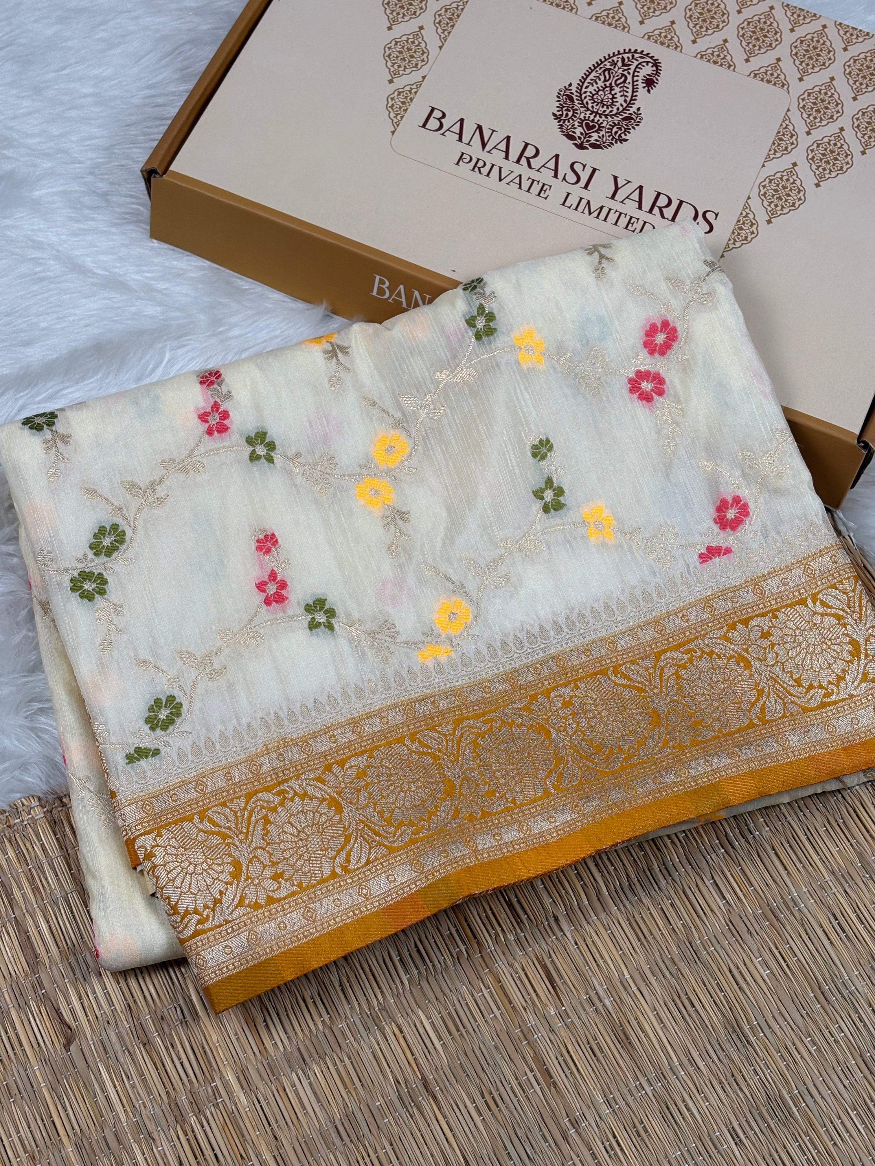 A folded white Banarasi silk saree with colorful floral mina weaving and a gold zari border, displayed next to its branded packaging box on a woven mat.