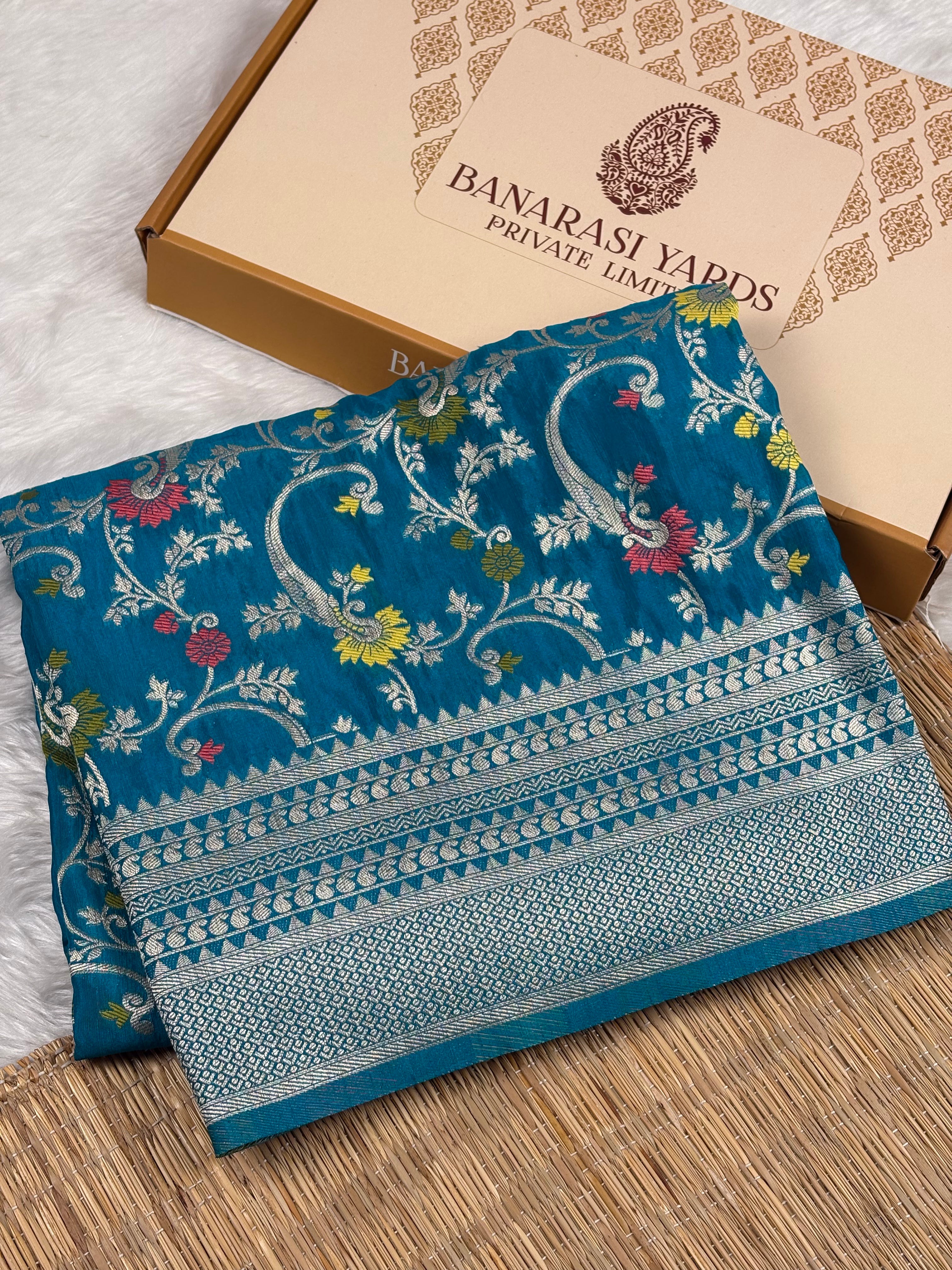 A folded teal blue Banarasi saree with intricate gold zari weaving and floral motifs, displayed next to its branded cardboard packaging box on a woven mat.