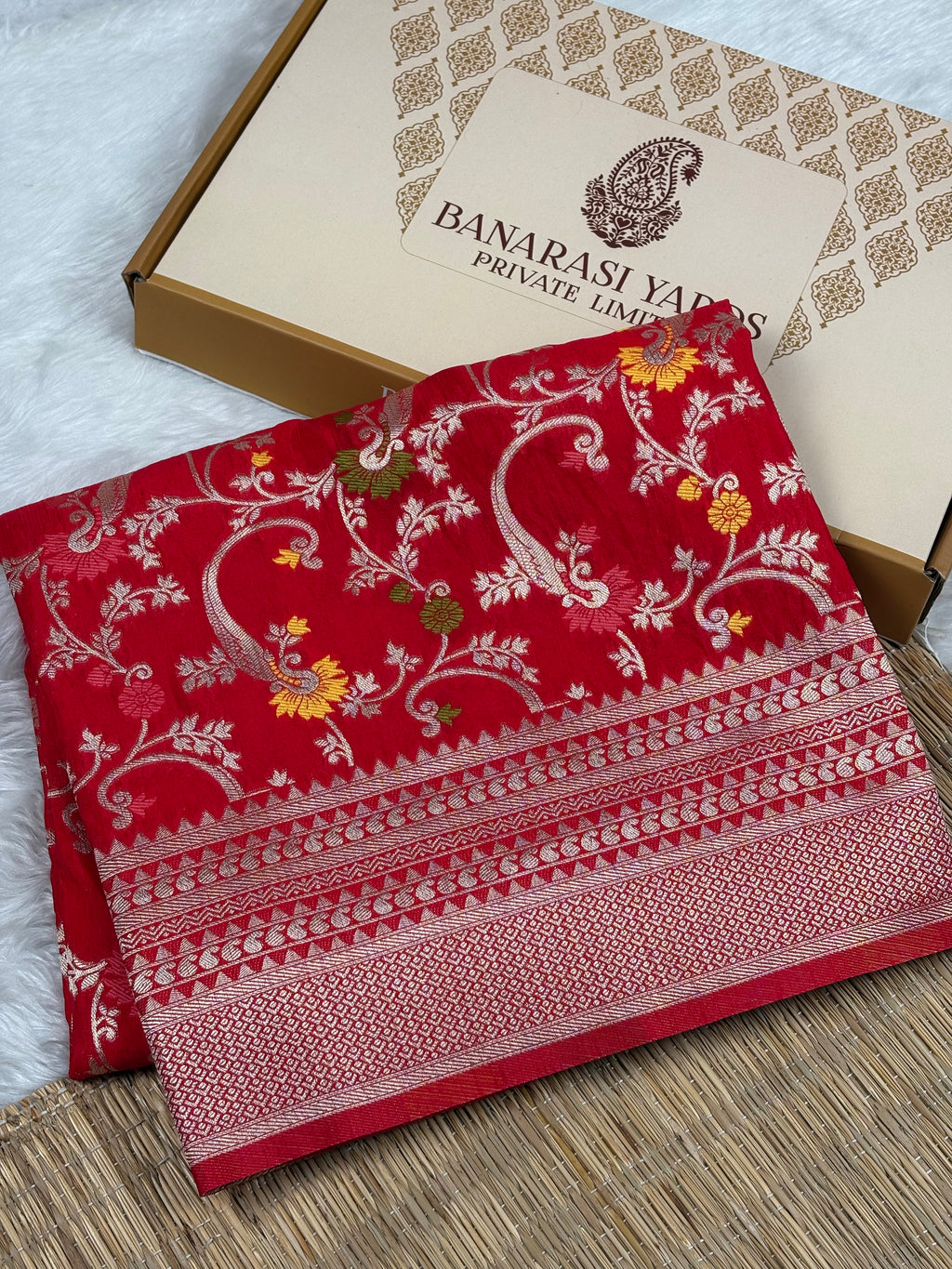 A folded red Banarasi silk saree with intricate gold zari weaving and floral mina motifs, displayed next to its beige presentation box labeled 'BANARASI YARDS PRIVATE LIMITED' on a woven mat.