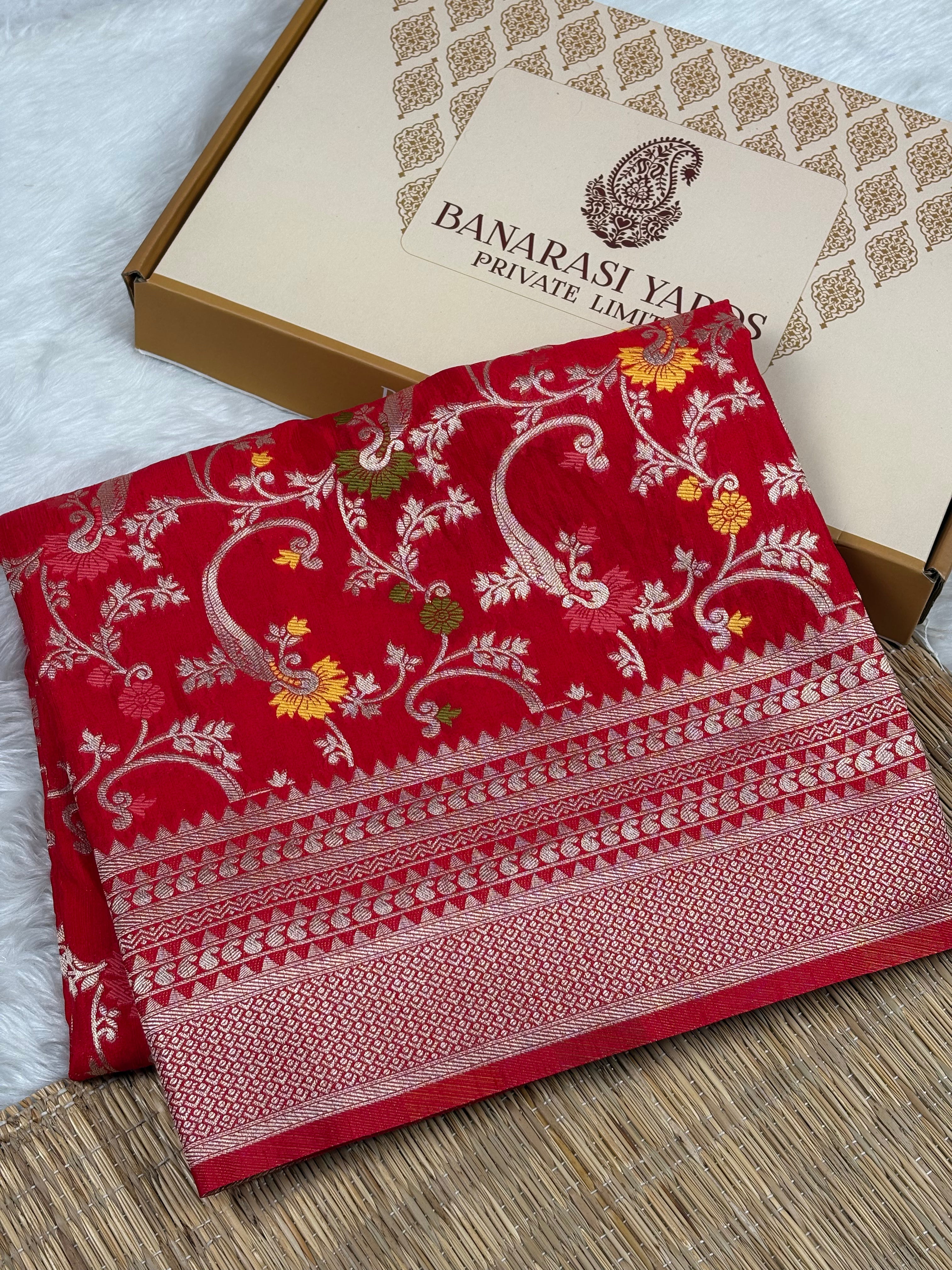 A folded red Banarasi silk saree with intricate gold zari weaving and floral mina motifs, displayed next to its beige presentation box labeled 'BANARASI YARDS PRIVATE LIMITED' on a woven mat.
