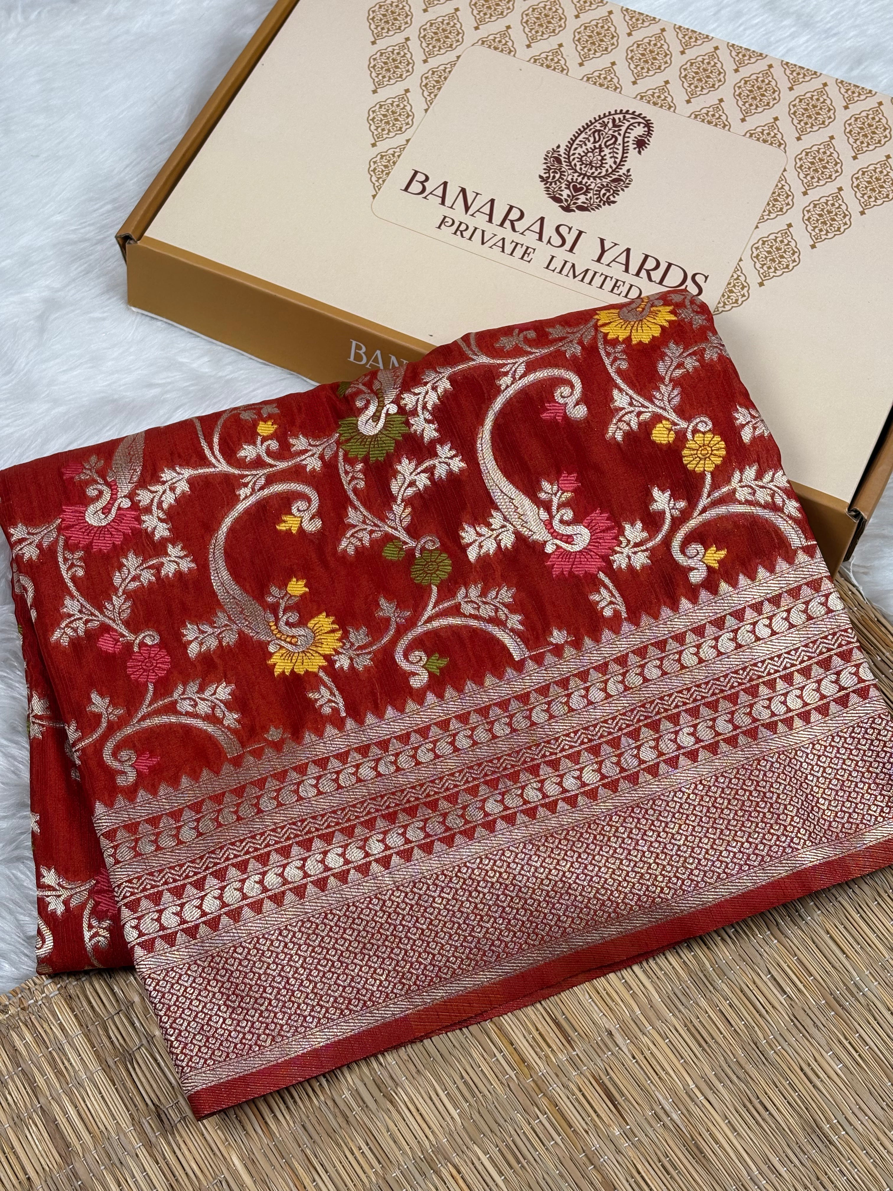 A folded red Banarasi silk saree with intricate yallow, pink, and green floral mina and zari weaving, displayed next to its beige Banarasi Yards Private Limited gift box on a woven mat.