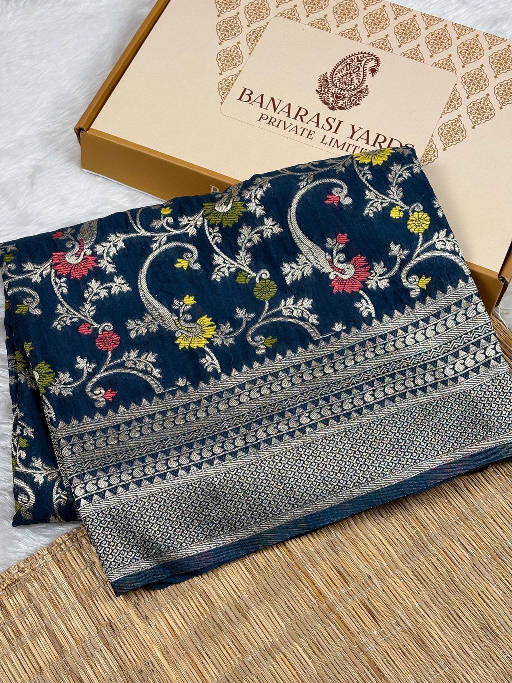 A folded navy blue Banarasi silk saree with intricate green, red, and yellow floral mina and zari weaving, displayed next to its beige branded packaging box on a woven mat.