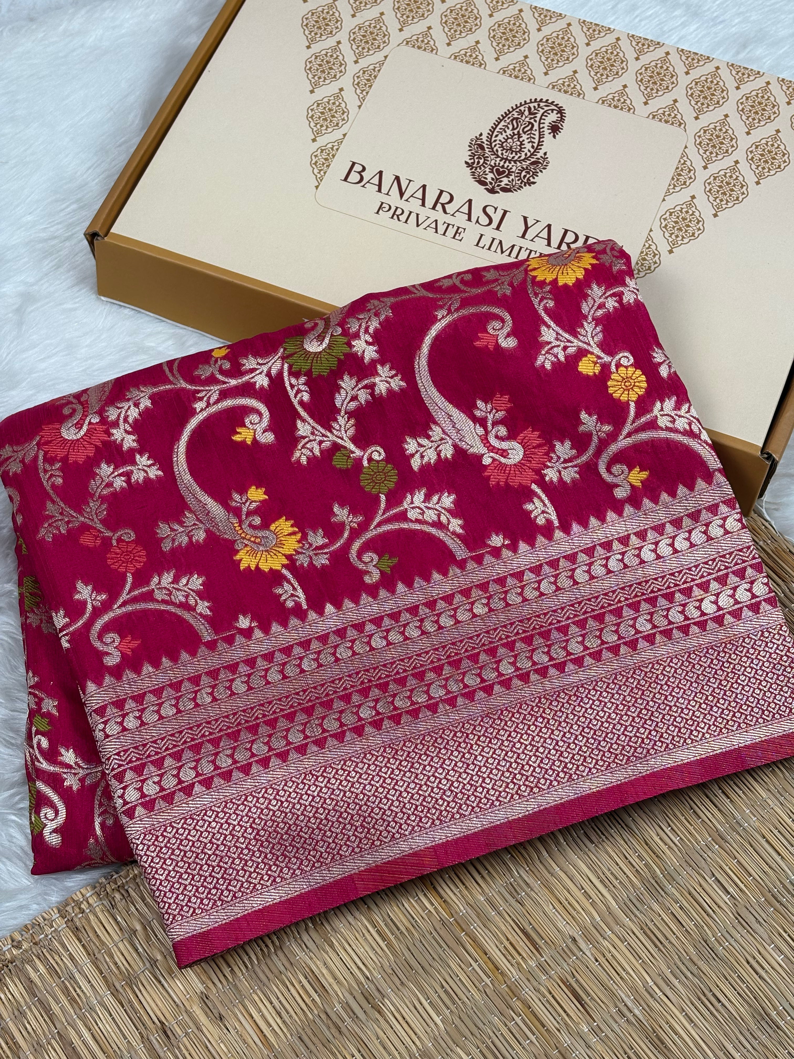 A folded deep pink Banarasi silk saree with intricate gold zari floral and paisley motifs, displayed next to its beige packaging box labeled 'BANARASI YARDS PRIVATE LIMITED' on a woven mat.