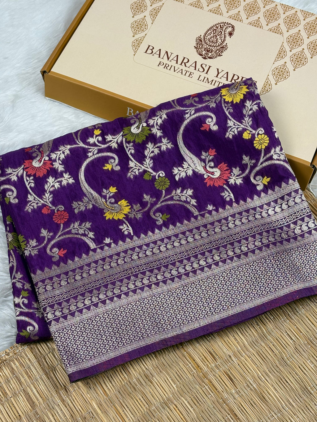 A folded purple Banarasi silk saree with intricate gold zari weaving and floral motifs, displayed next to its branded packaging box on a woven mat.