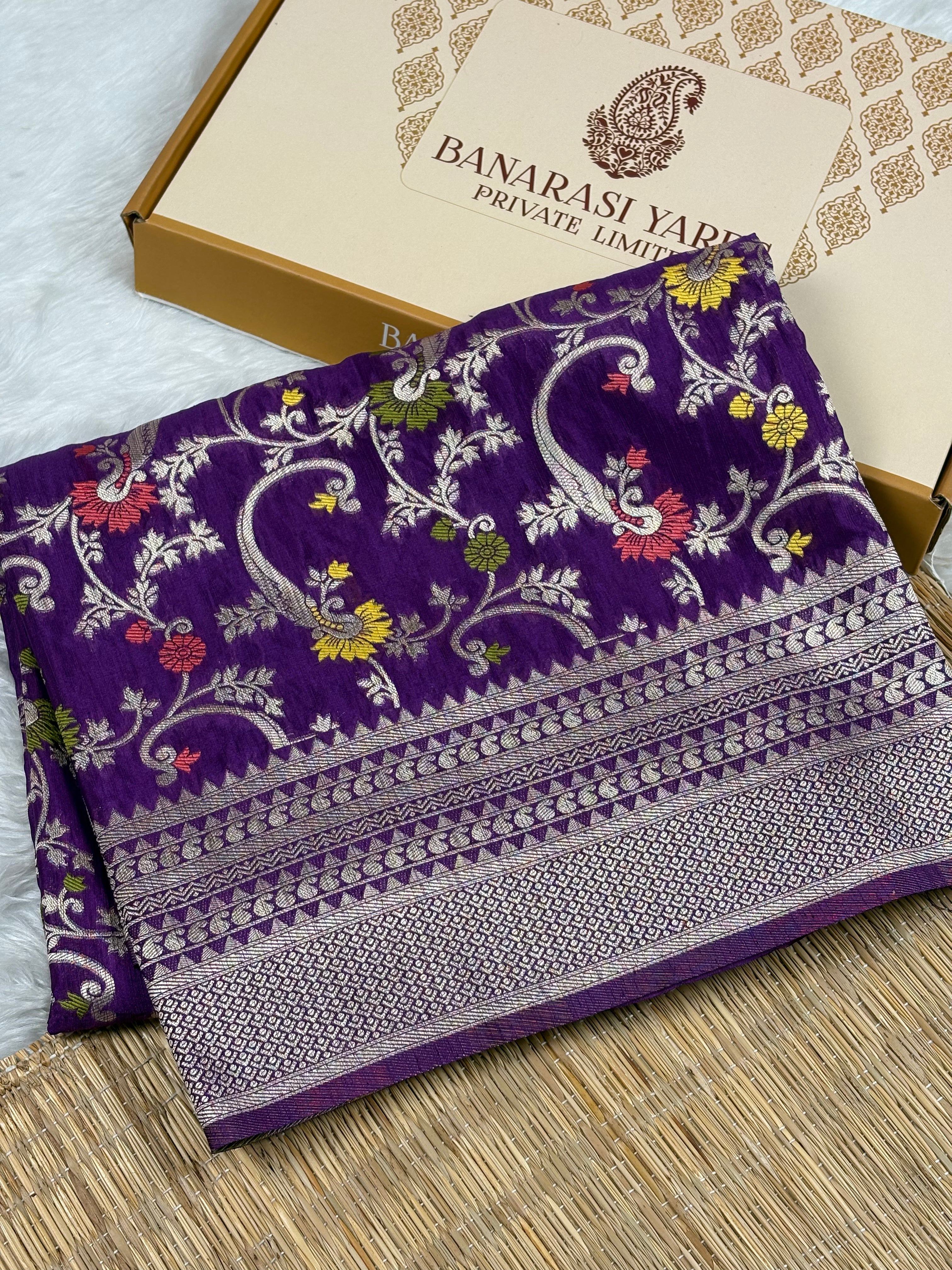 A folded purple Banarasi silk saree with intricate gold zari weaving and floral motifs, displayed next to its branded packaging box on a woven mat.