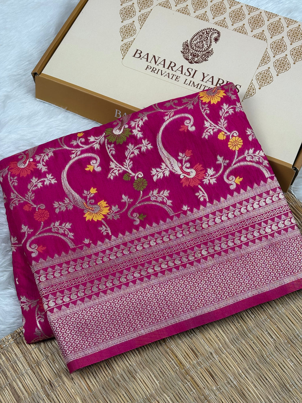 A folded magenta pink Banarasi silk saree with intricate gold zari weaving, displayed next to its branded gift box on a woven mat.