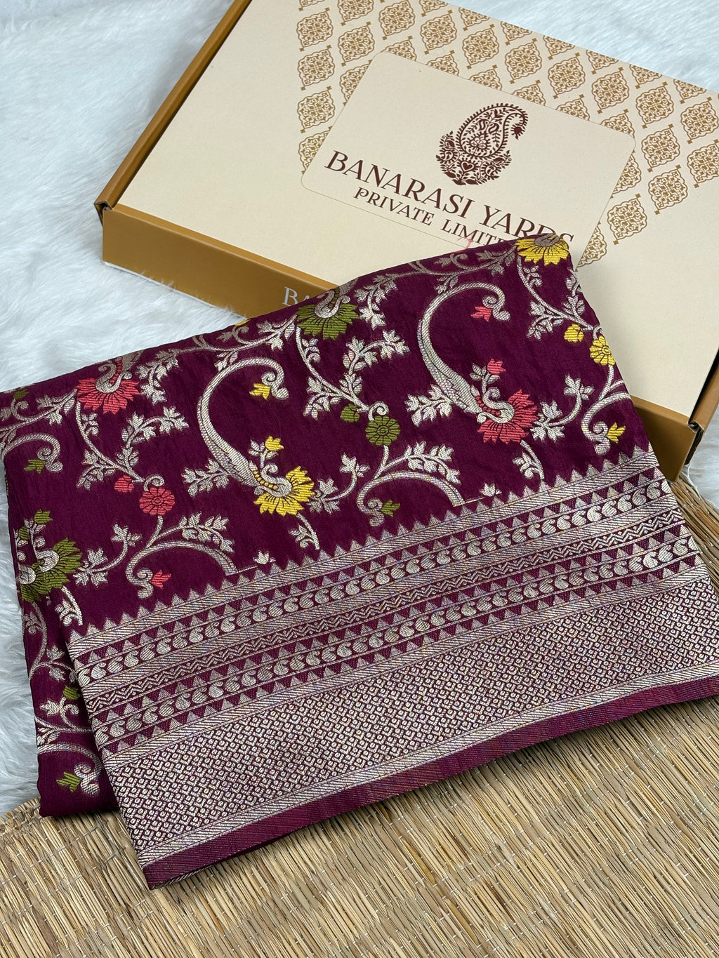 A folded deep purple Banarasi silk saree with intricate floral and paisley zari Weaving, displayed next to its branded packaging box on a woven mat.