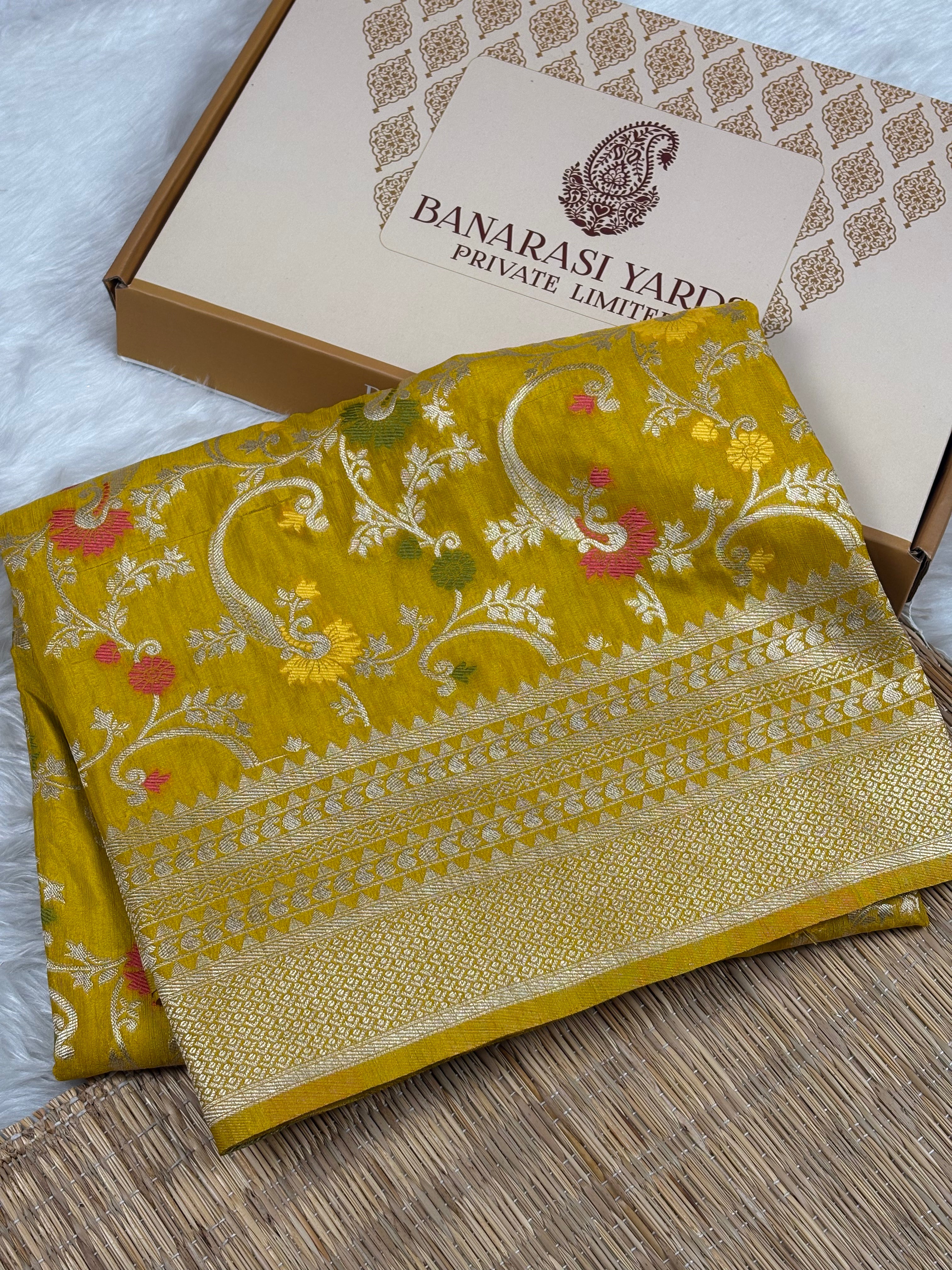 A folded mustard yellow Banarasi silk saree with intricate floral mina and paisley zari weaving, displayed next to its branded cardboard box on a woven mat.