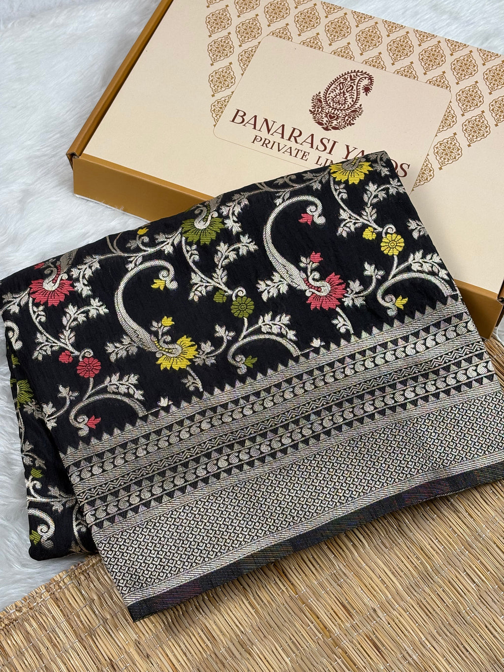 A folded black Banarasi silk saree with intricate floral and paisley zari weaving, displayed next to its beige packaging box on a neutral background.