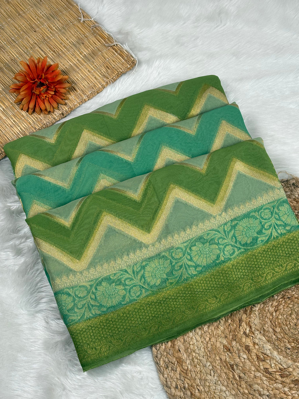 A folded green and teal chevron-patterned saree with a floral border, displayed on a white faux fur surface with decorative woven mats and an orange flower.