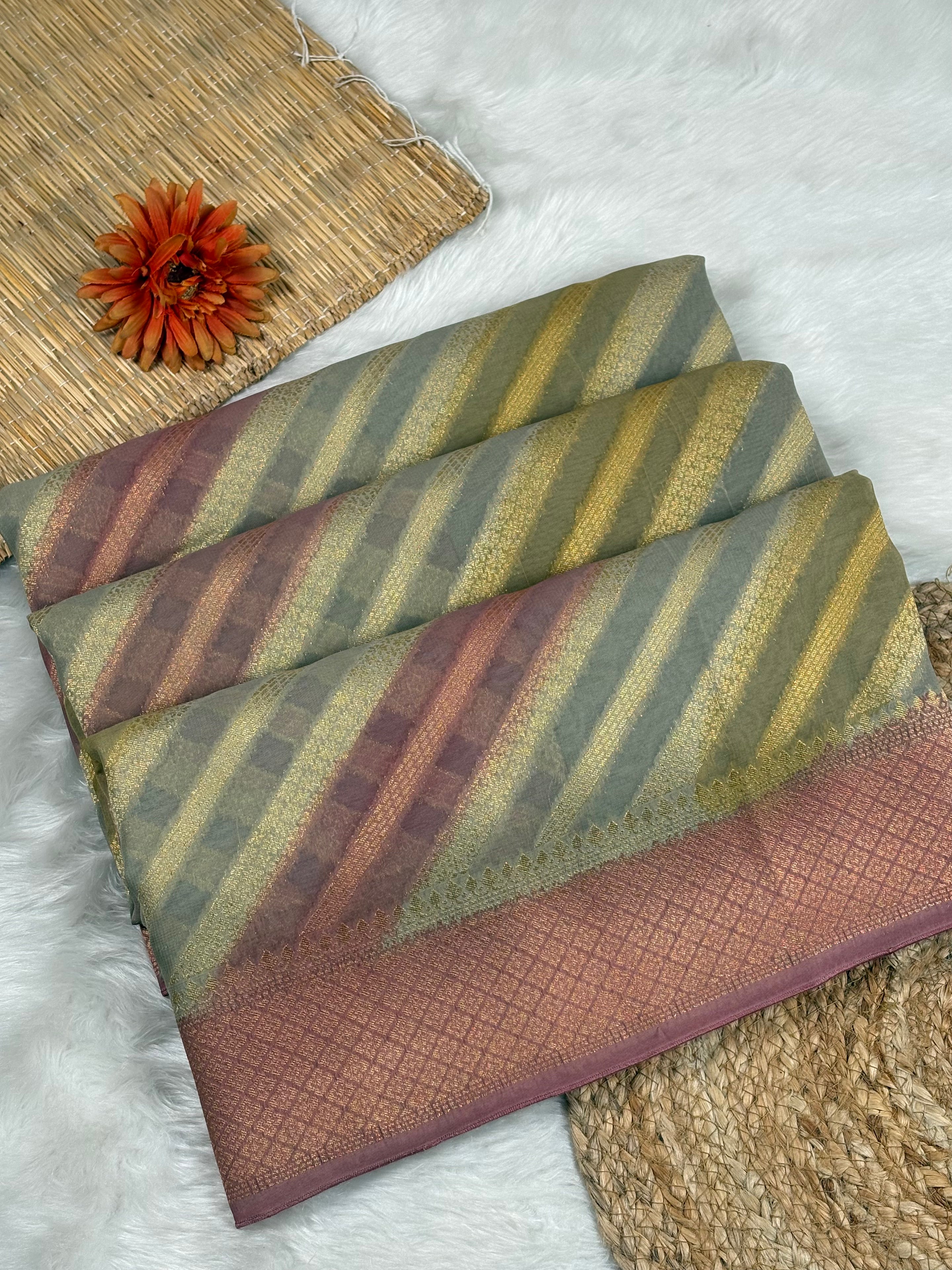 A folded striped fabric piece, saree, displayed on a white faux fur surface with decorative props including a woven mat, a dried orange flower, and a jute runner. The fabric features diagonal stripes in muted olive green, mustard yellow, dusty rose, and brown, with a solid patterned border in dusty rose.