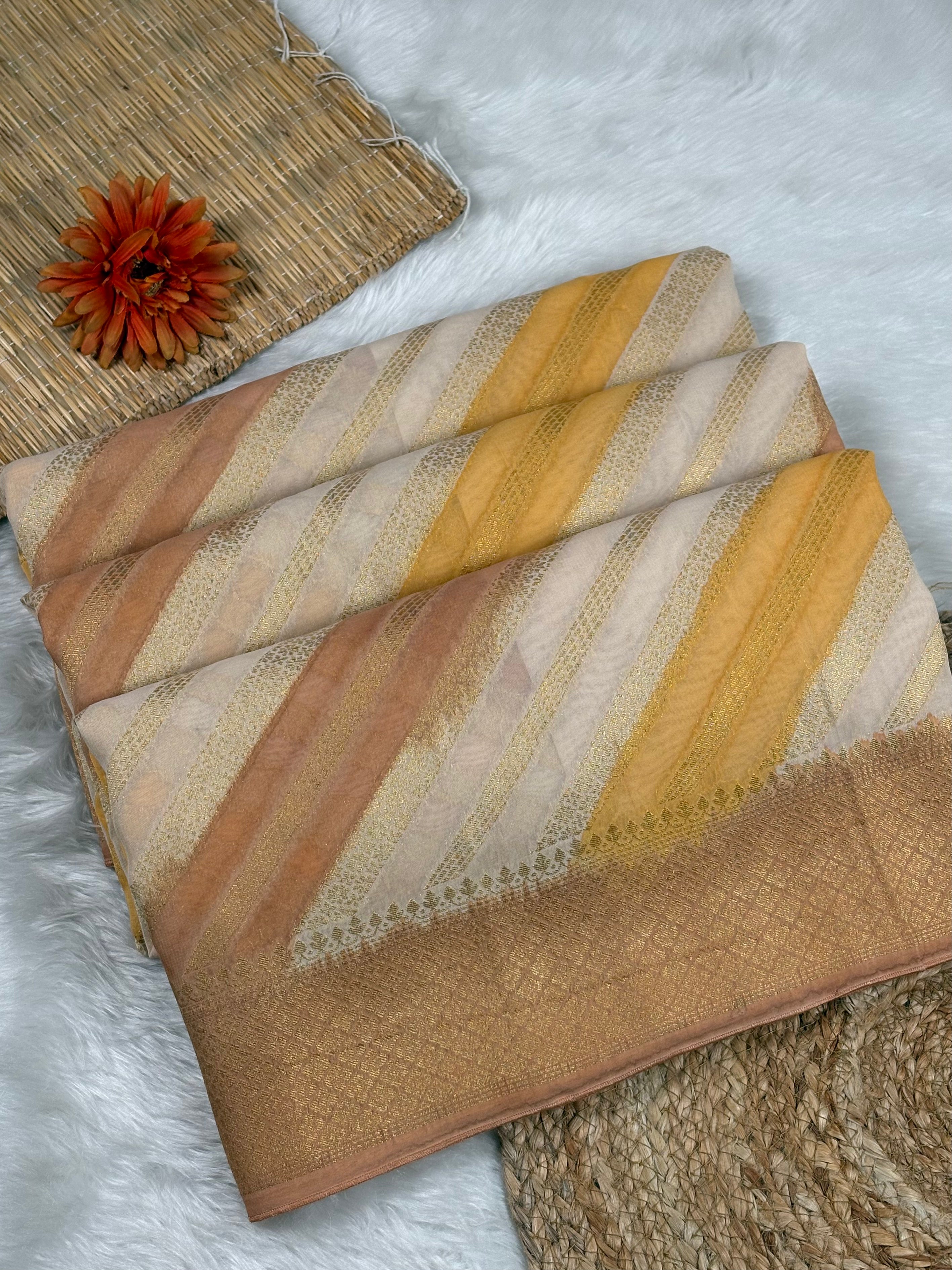A folded striped saree in warm beige, cream, tan, and mustard yellow tones, styled on a white faux-fur surface with woven mats and a small orange flower prop.