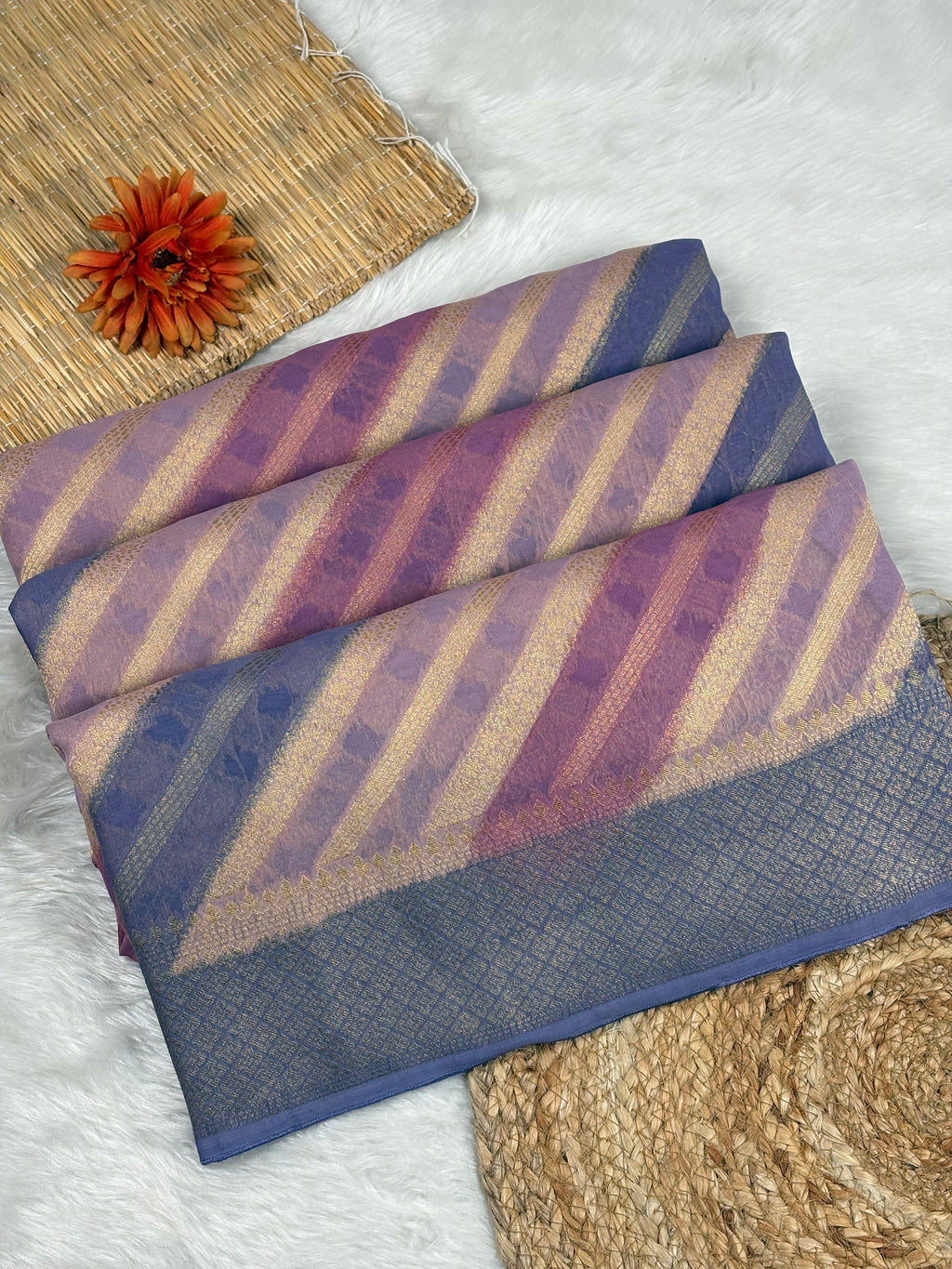 A folded multicolor striped saree with purple, blue, and beige diagonal bands and a blue border, styled on a white faux fur surface with woven mats and an orange flower.