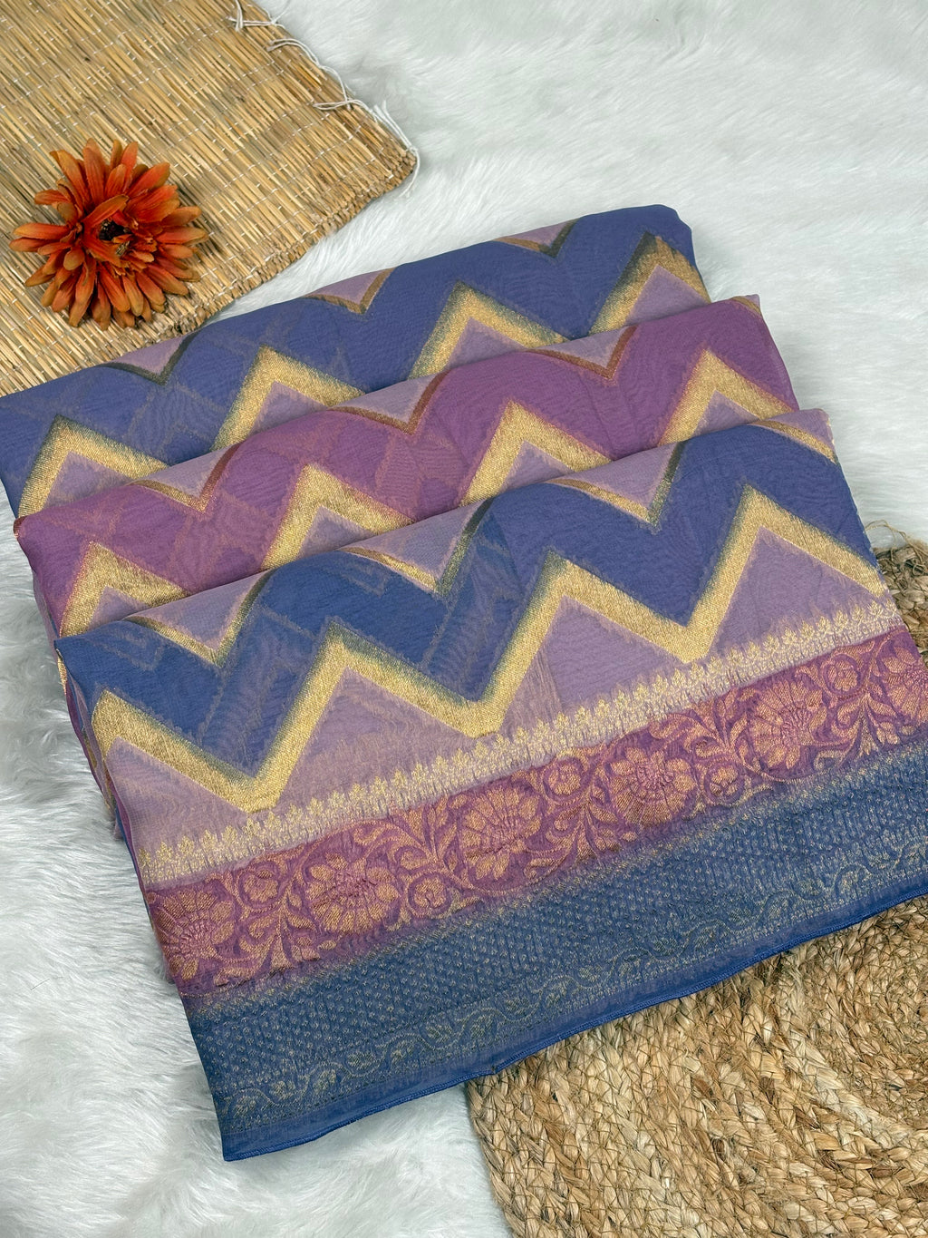 A folded patterned fabric saree with purple and blue color, gold zigzag zari stripes and a floral border, styled on a white faux-fur surface with woven mat props and a small orange flower.